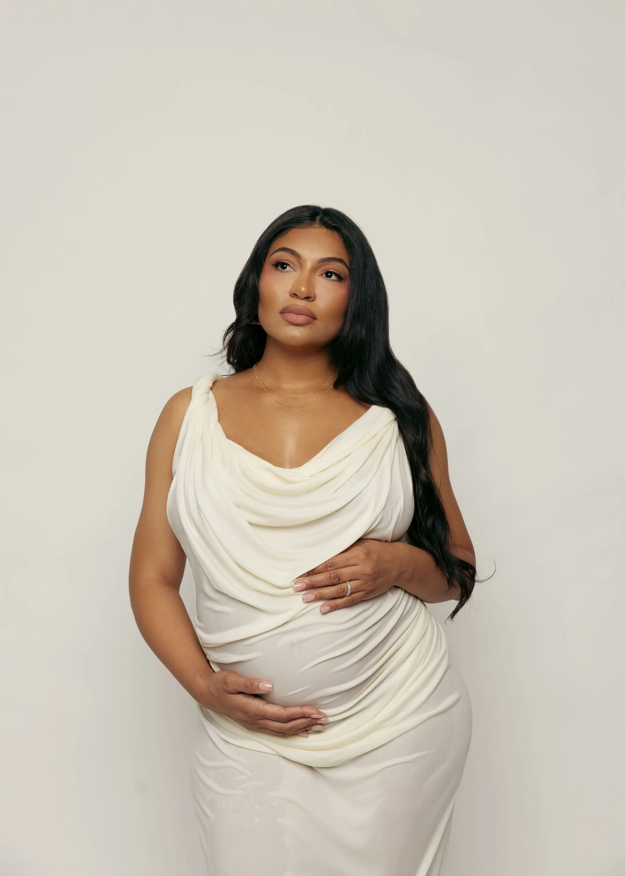 Pregnant woman with long dark hair wearing a cream dress, standing against a plain light background, gently holding her belly with one hand and looking thoughtfully.
