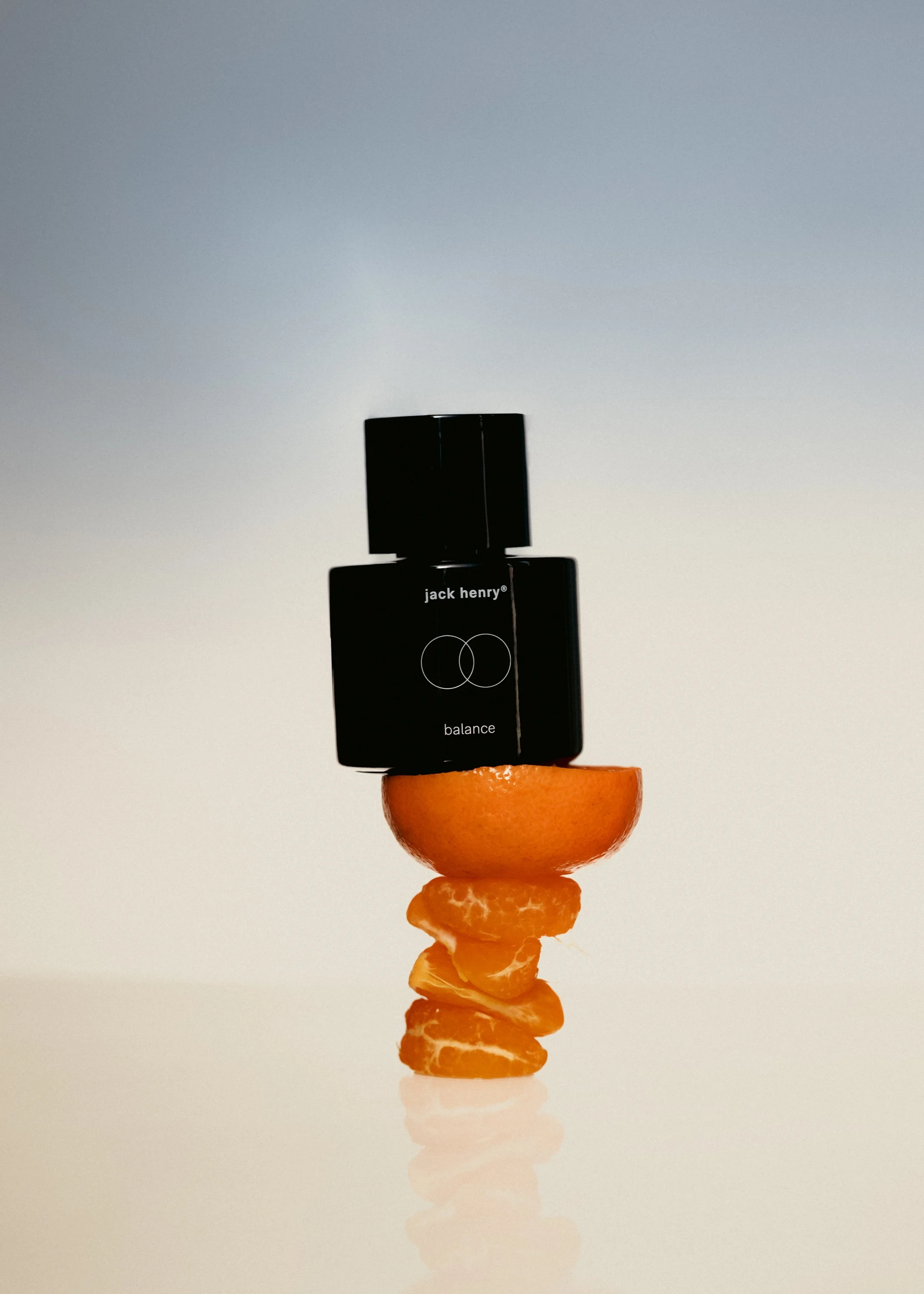 A black perfume bottle labeled 'jack henry' and 'balance' balanced on a half-cut orange, with orange peels stacked below it, set against a plain background.