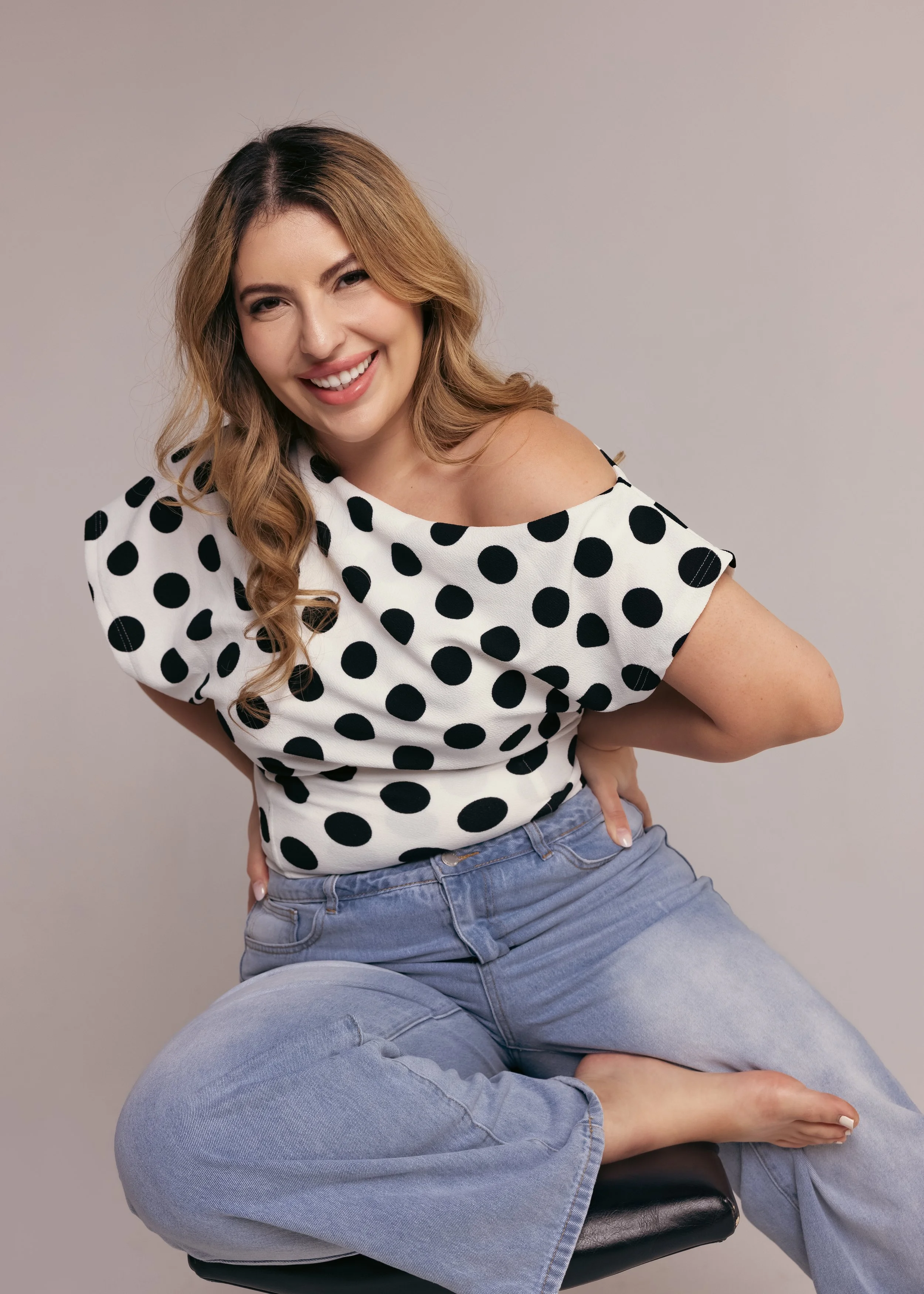 A woman with blond hair wearing a white top with black polka dots, sitting cross-legged on a stool, smiling at the camera.