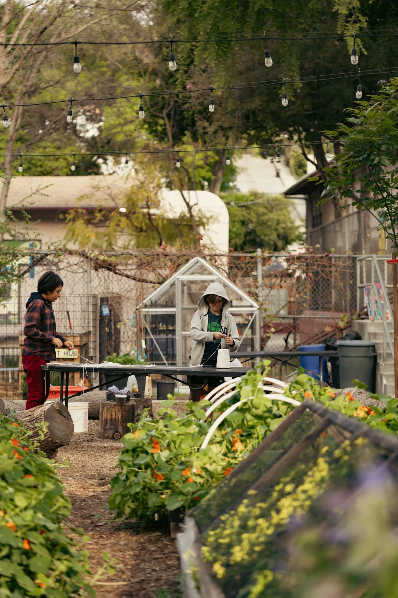 Two children in a community garden, with one working at a table and the other tending to plants, surrounded by green plants, trees, and string lights overhead.