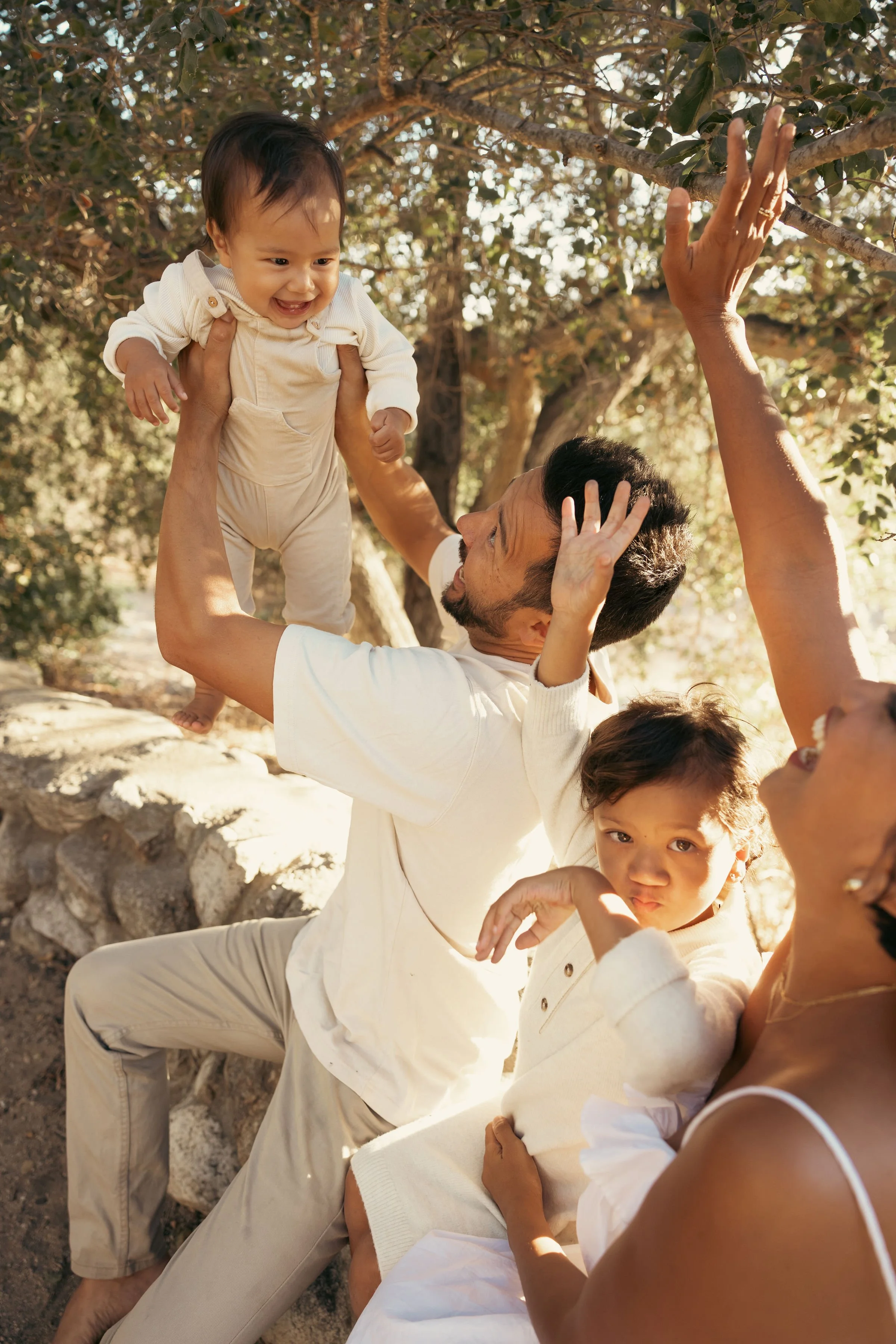 A man lifting a smiling baby in a cream outfit in an outdoor setting, with a woman and a young girl nearby, all under a tree with dappled sunlight.