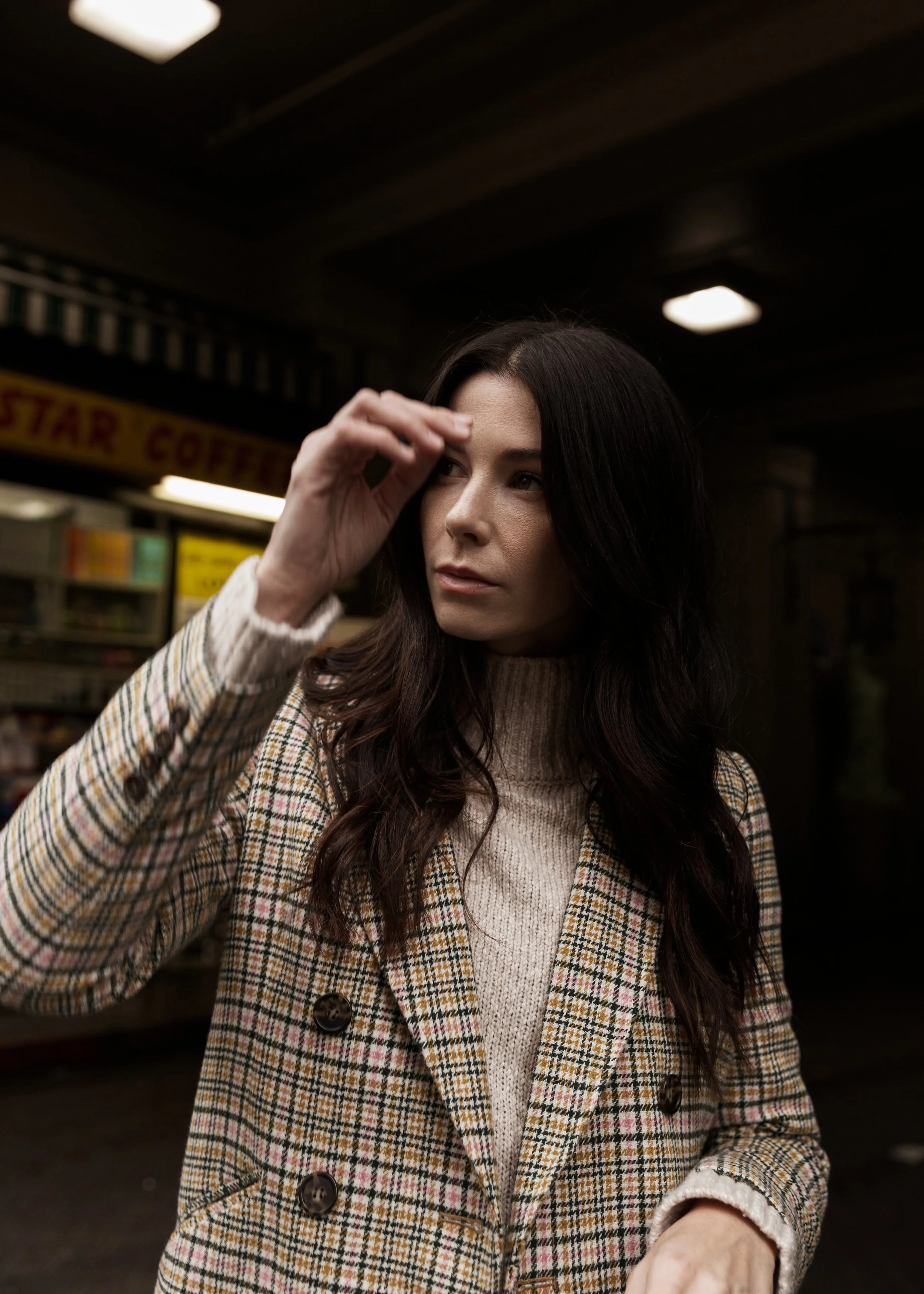 A woman with dark hair wearing a plaid blazer and a turtleneck sweater, standing indoors with a dark background and a sign that partially reads "STAR COFFEE."