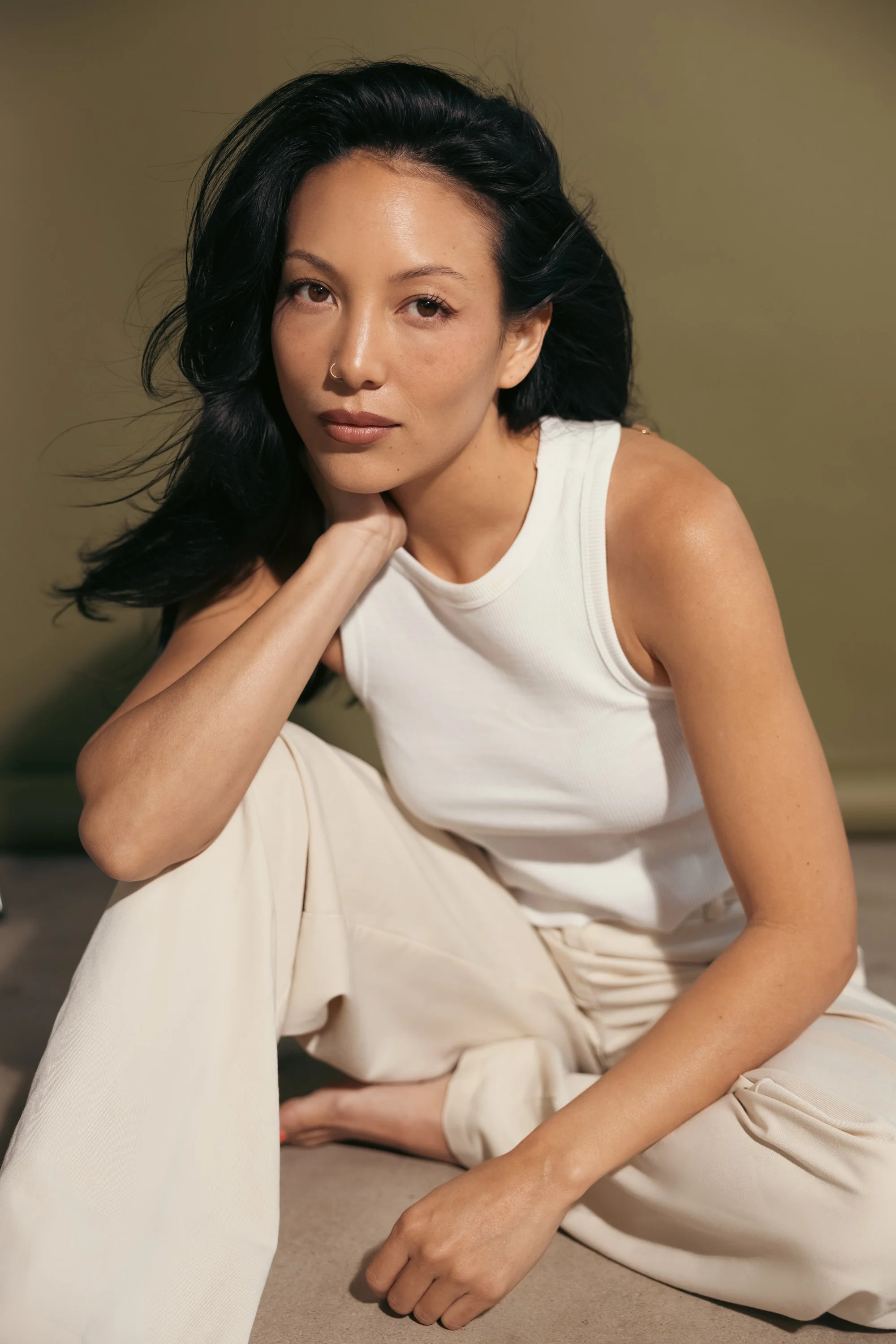 A woman with black hair, wearing a white sleeveless top and cream-colored pants, sitting on the floor with her leg bent and her chin resting on her hand, looking into the camera.