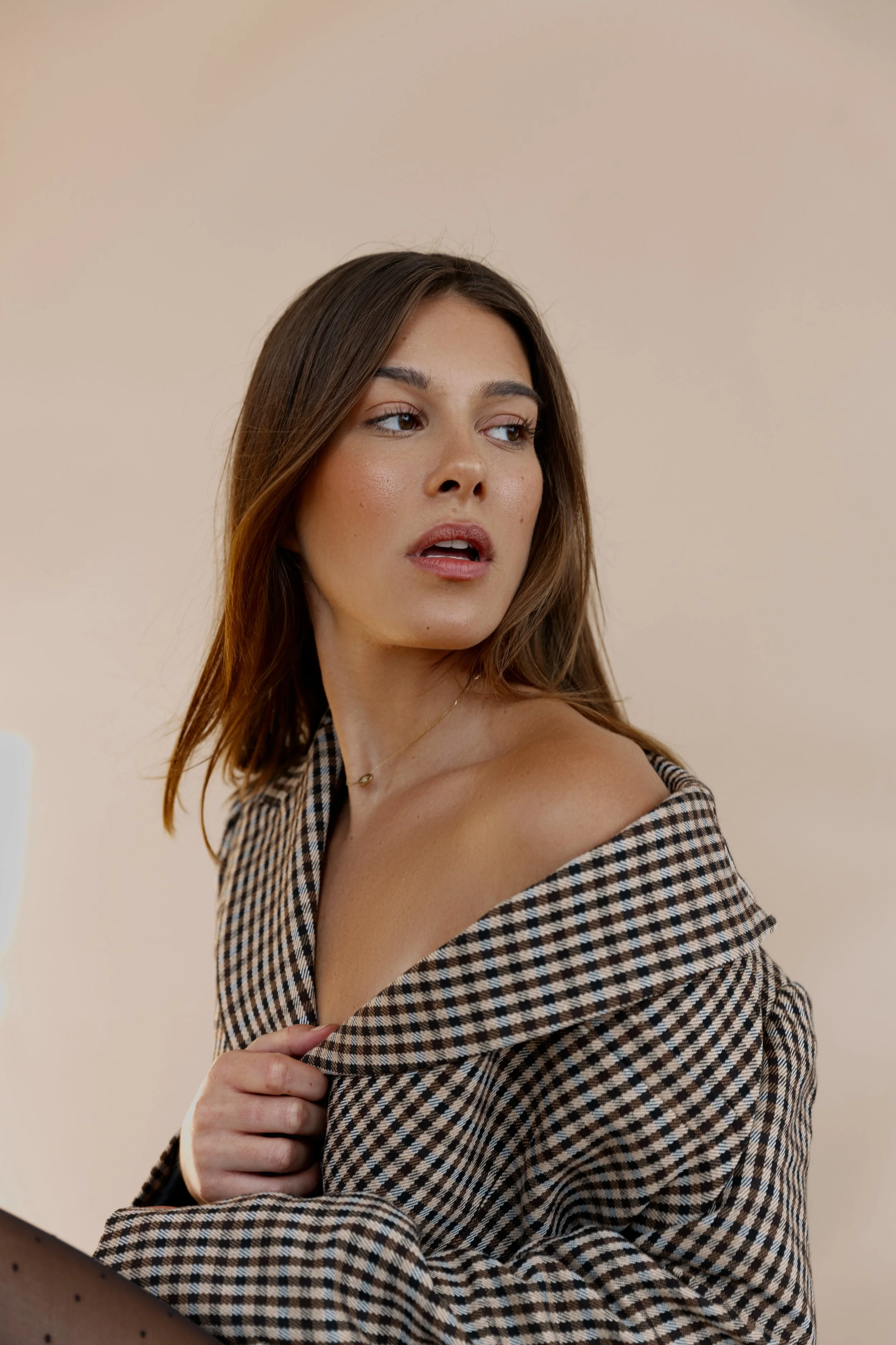 A woman with shoulder-length brown hair, wearing a checkered off-the-shoulder top, looking to the side with a neutral expression.