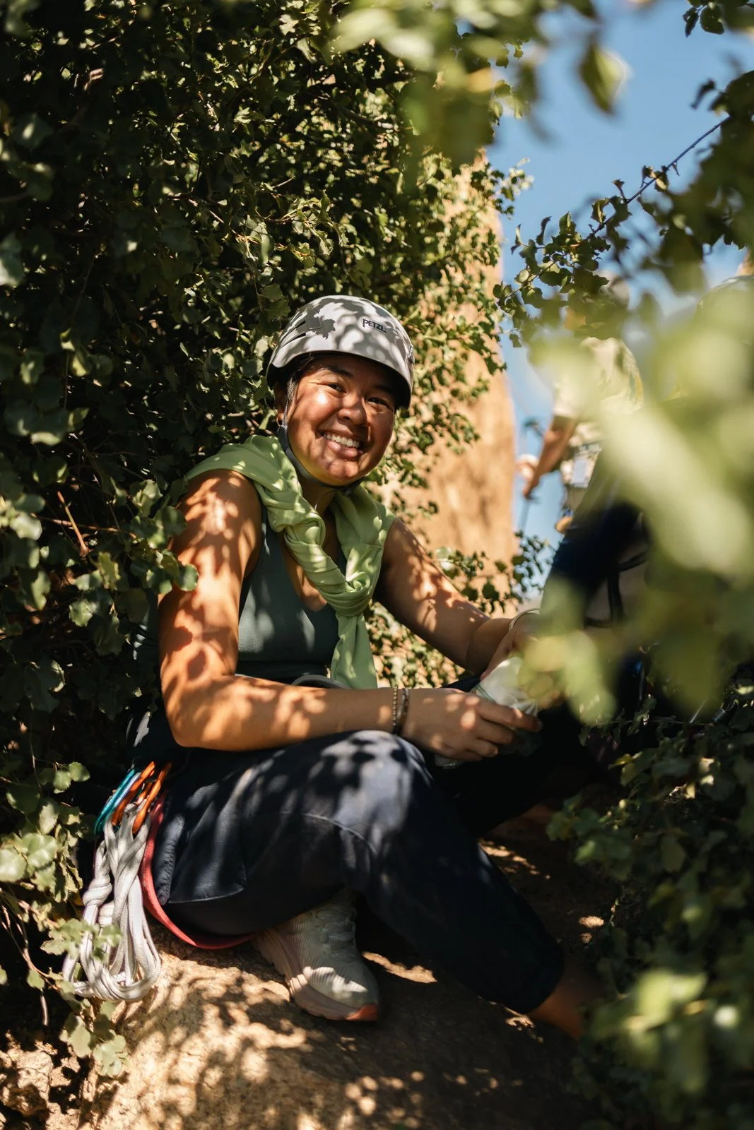 Person smiling while sitting between bushes outdoors, wearing a helmet, with climbing gear attached to pants.