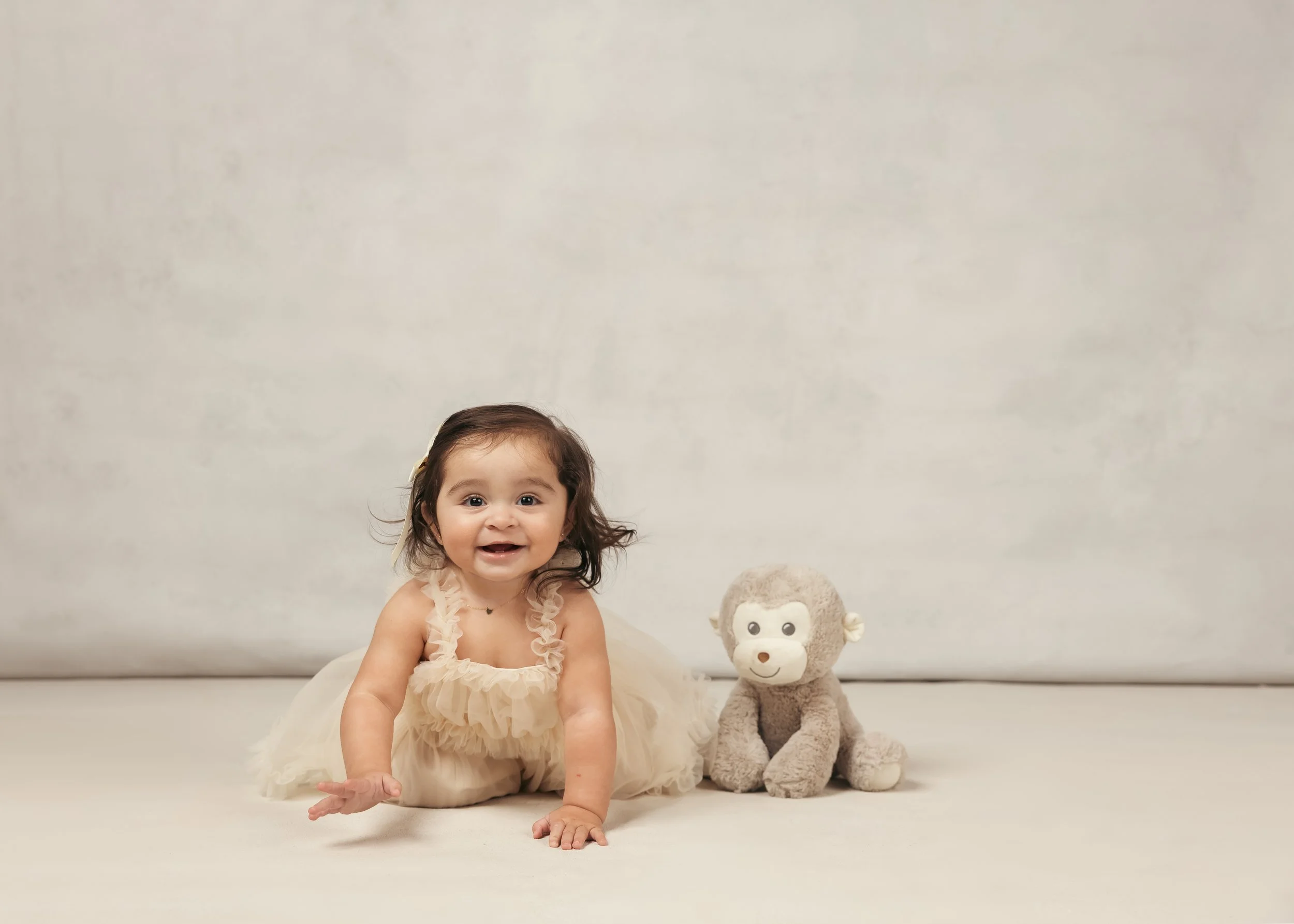 A smiling baby girl with dark hair lying on her stomach next to a plush monkey toy against a plain background.