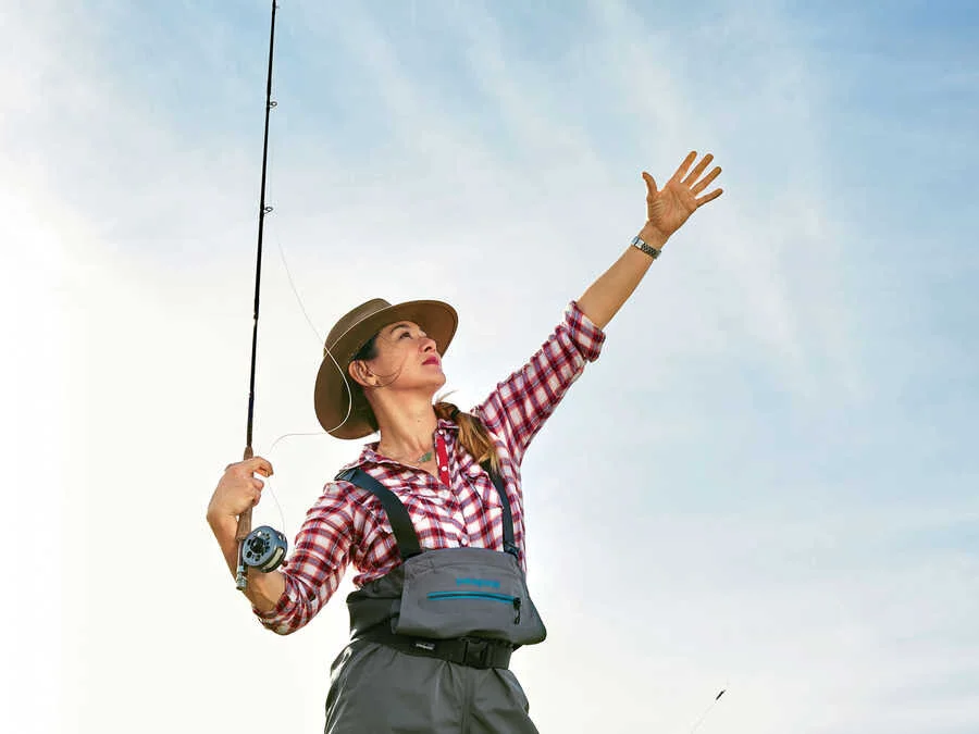 Author Kim Cross fly fishing, reaching for the line against a backlit sky
