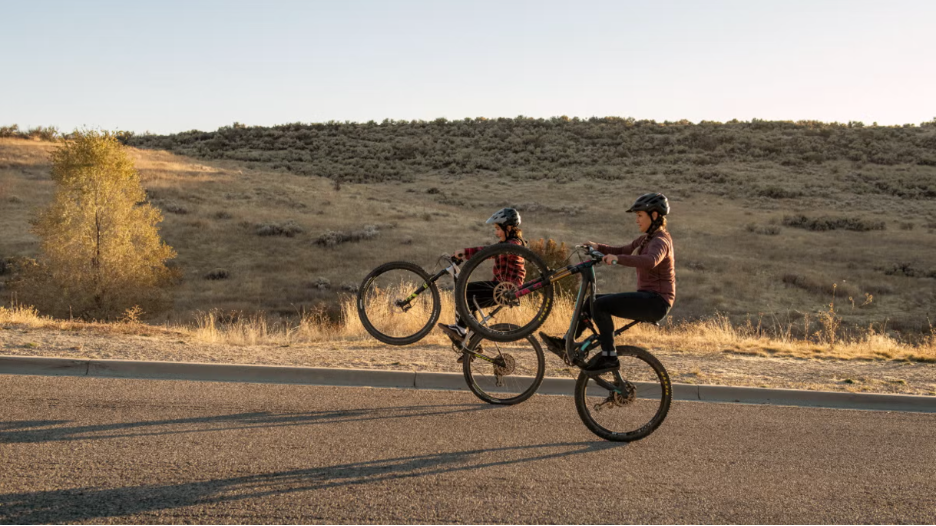 A mother and young son practice wheelies side by side on mountain bikes with open space in the background
