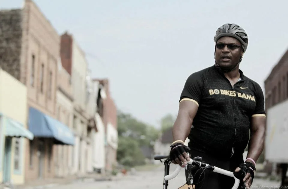 Bo Jackson, ESPN's Greatest Athlete of All Time, wearing a helmet and cycling jersey astride a road bike with a small tornado-torn town in the background 