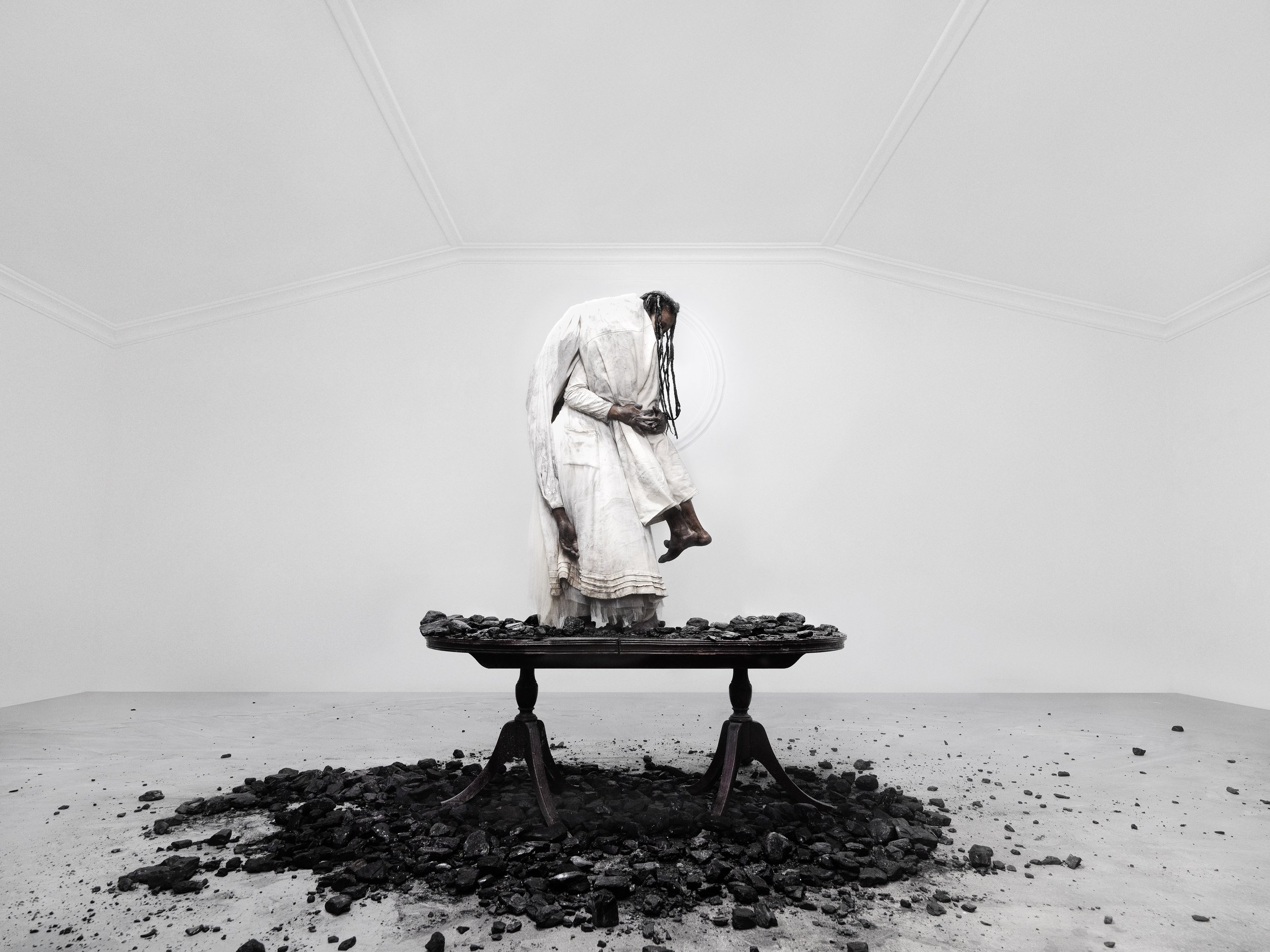 ZION deals with the relationship between body and place. The idea that all people are meant to belong somewhere, some place, yet in reality there are millions of people that are unsettled, in search of refuge, migrating across borders and landscapes for various reasons. In this work there is no obvious signifier of place except for a heap of coal being thrown on a table, onto the floor and over he body. The black rocks of fossilised material represents the abundant natural mineral wealth deposited beneath the ground. Whilst coal is a valuable source of energy across the world, sustaining modern economies and industries, the more valued minerals such as oil, gold, copper and diamonds, which are found in abundance on the African continent, often breed and fuel conflict. And whilst most people are forced to flee from their homes due to harsh climate and collapsing economies, civil war is amongst the leading causes of migration across the african continent. As such, the depiction of coal in this series not only refers to the mineral wealth attached to place, it also refers to the absence of place as people flee from their homes due to the violence that follows the exploitation of these types of mineral resources. The Zion series thus refers not only to the absence of place/home but also to the many conflicts that have caused the forced migration of millions of African people within the African context, casting them into a life of refuge. The series depicts a struggle of a woman, who in the time of conflict often assumes the role of the provider and a carer, struggles to retain some sense of home....she is seen at the diner table serving a resource for the (human) family to consume...while in a fleeting state. In this work the tensions of this relationship of exploitation is anchored by an unusual love story.