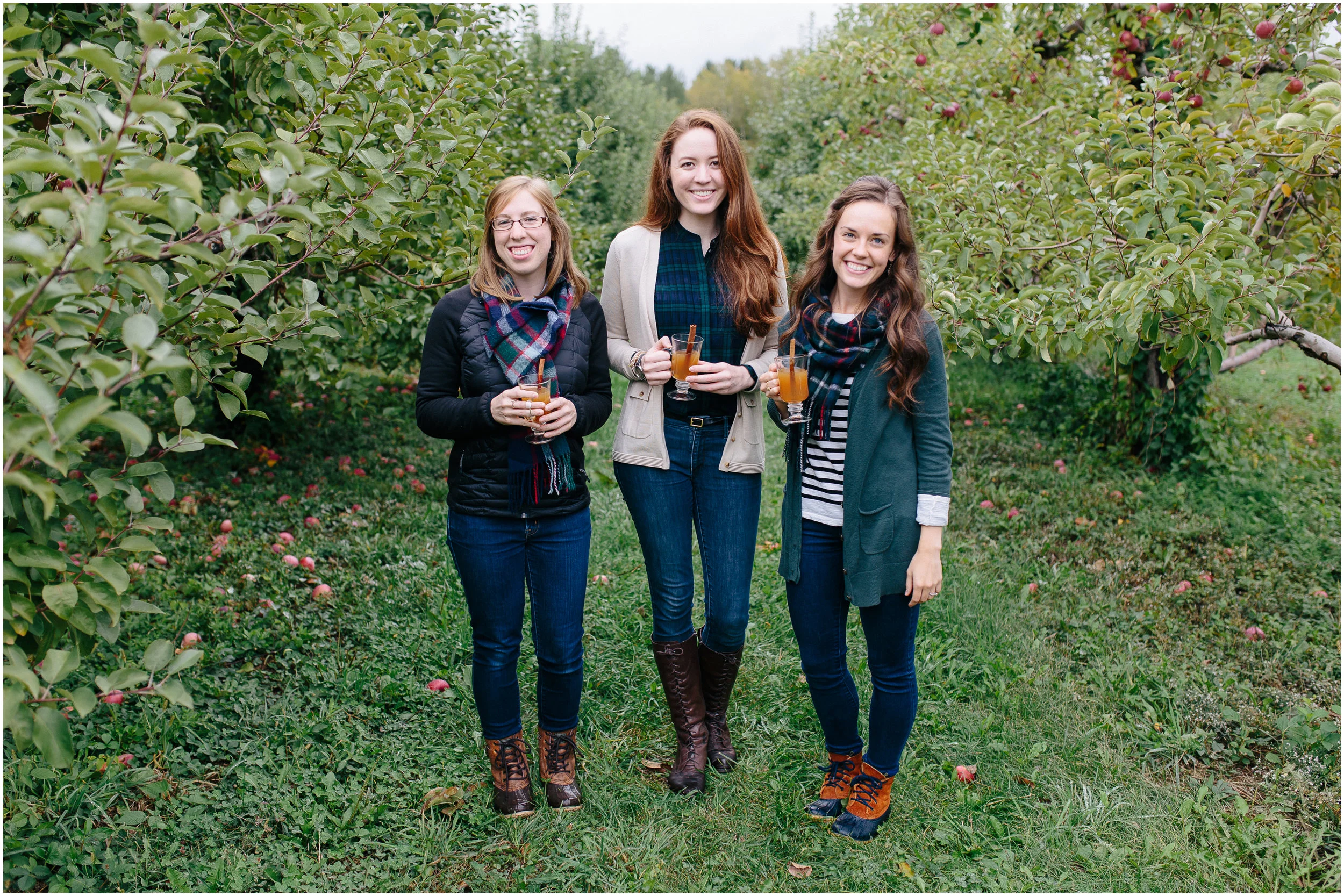Girl friends smiling in an apple orchard, holding hot apple cider with cinnamon sticks, cute scarves, trendy