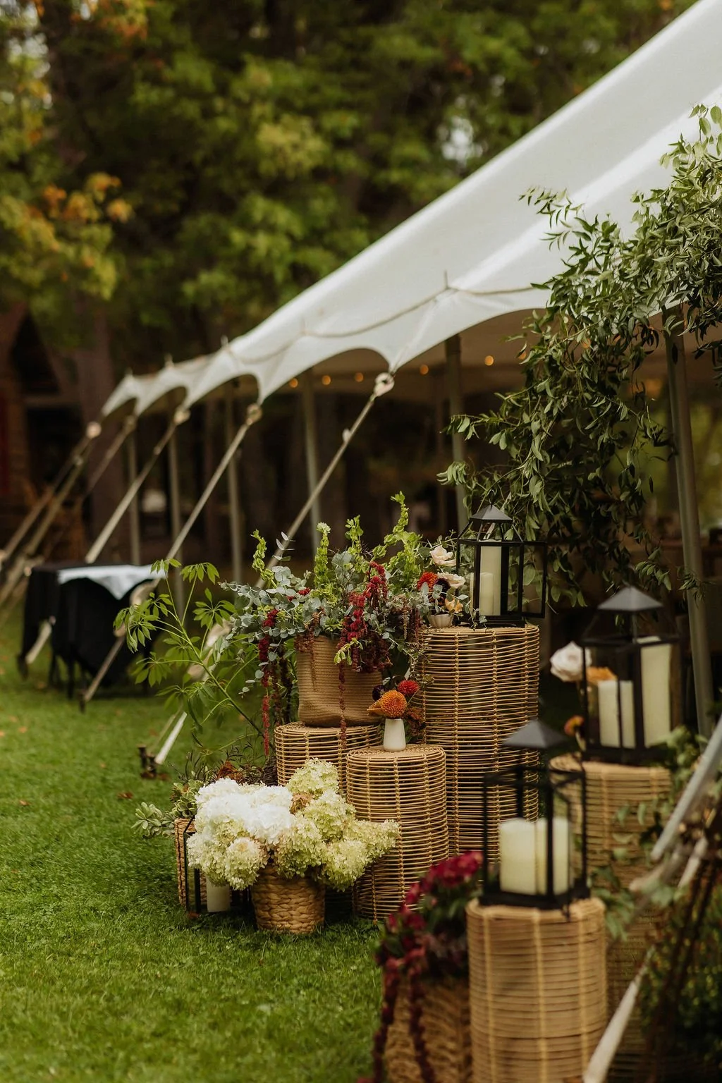 The image depicts an outdoor wedding or event setup under a white canopy tent. The decor includes wicker pedestals with floral arrangements, candles in lanterns, and lush green foliage, creating an elegant and natural ambiance.