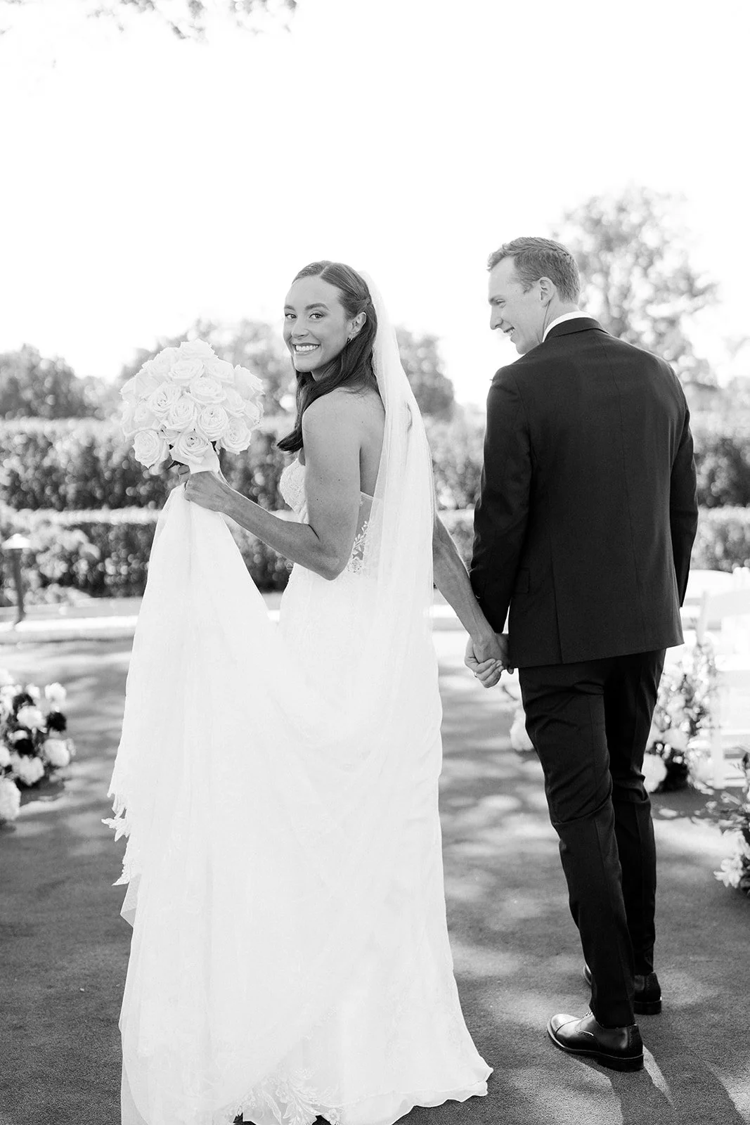 A smiling bride holding a bouquet of roses, holding hands with her groom during their wedding ceremony outdoors. Minekahda club maroon mulberry florals wedding planning minneapolis wedding planner 