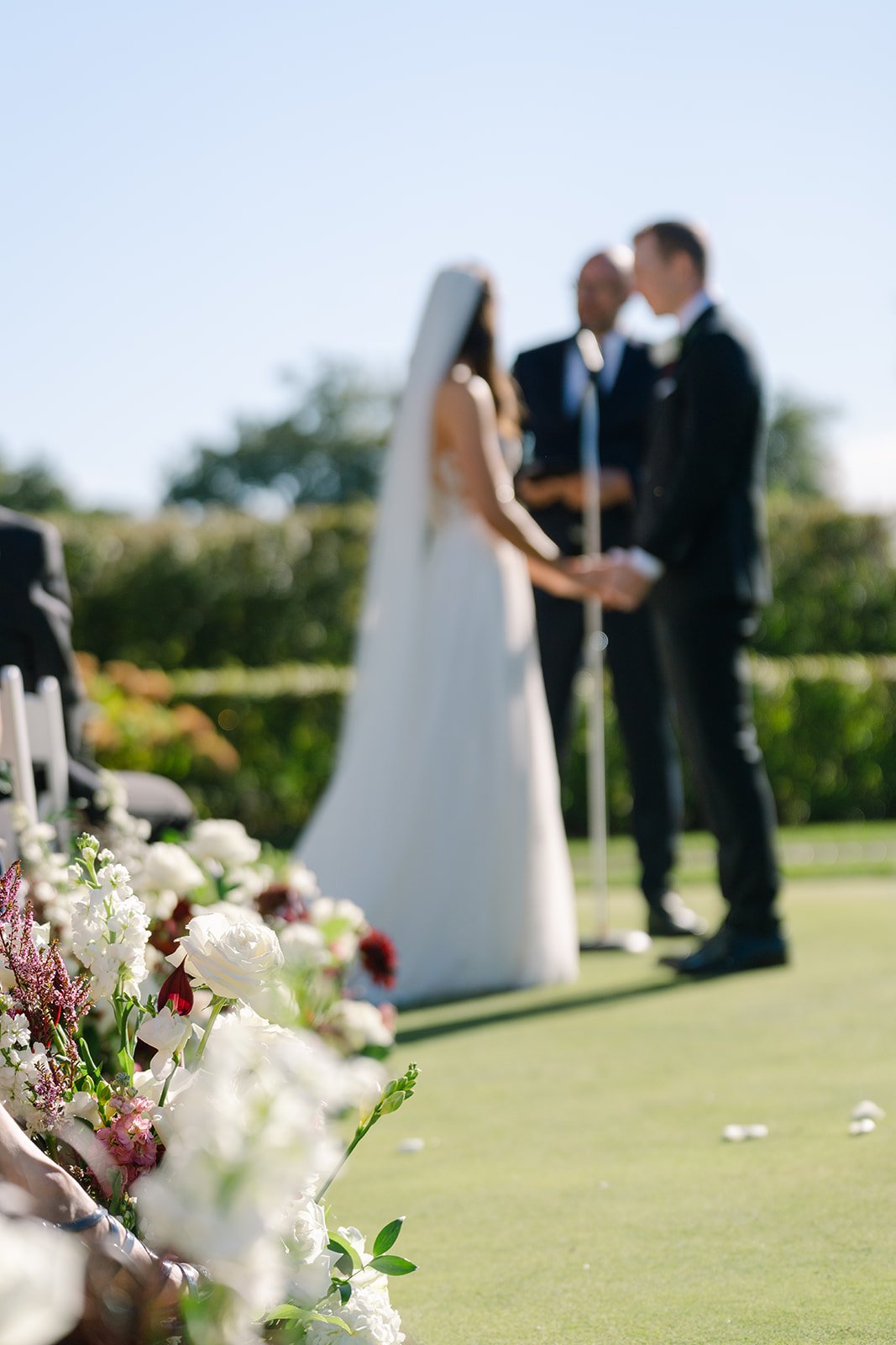 A blurred photo of a wedding ceremony outdoors with a bride and groom holding hands in front of an officiant, with floral arrangements in the foreground.