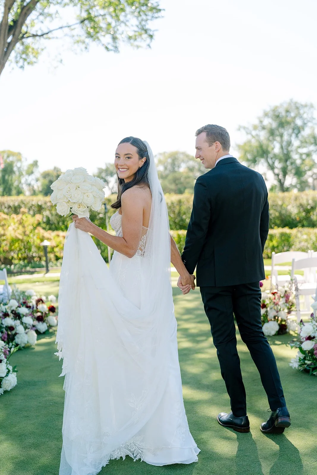 A bride in a white wedding dress holding a bouquet of white roses, smiling at the camera, and a groom in a black suit holding her hand, standing outdoors on a sunny day with flowers and greenery around.