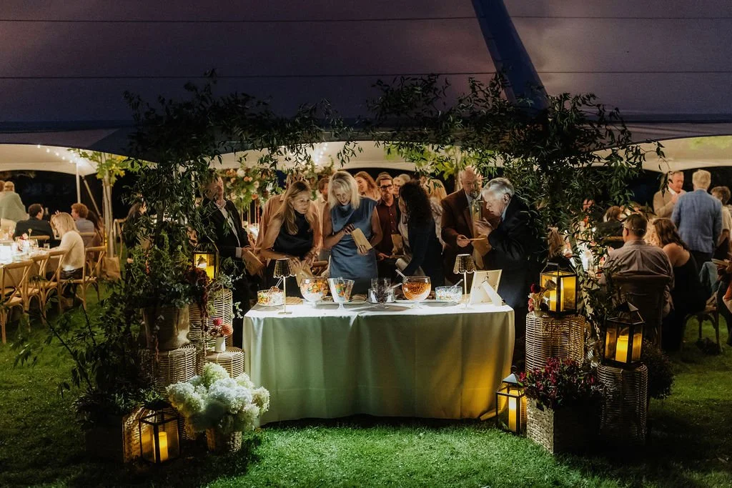 People gather around a table with fish bowls and lamps under a canopy at a formal outdoor event, with other guests seated at tables in the background.
