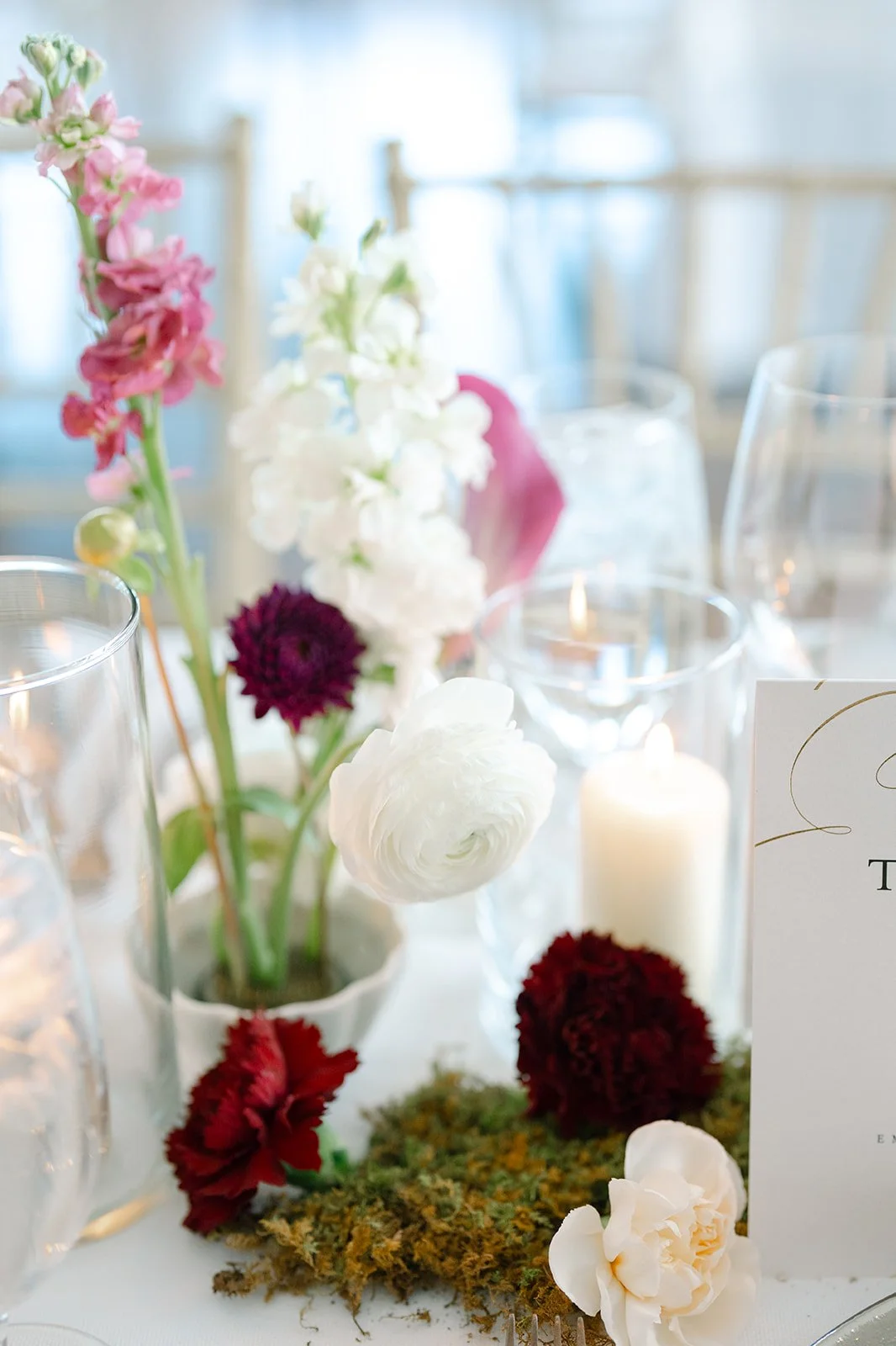 A floral centerpiece with pink, white, and dark purple flowers on a table, surrounded by candles and a white place card.