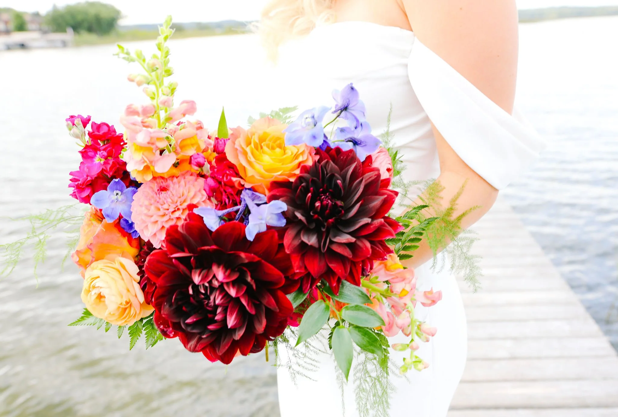 Woman in a white dress holding a colorful bouquet of flowers by the water on a dock. Maddens wedding resort florist