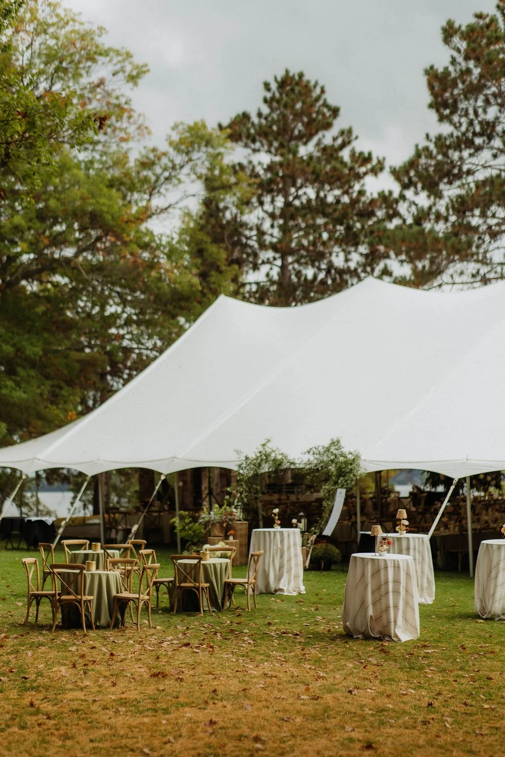 Outdoor event setup with round tables, chairs, and high-top tables under a large white canopy tent on a grassy area, surrounded by trees. tent wedding  camp foley wedding planning floral northern minnesota wedding jenkins nisswa