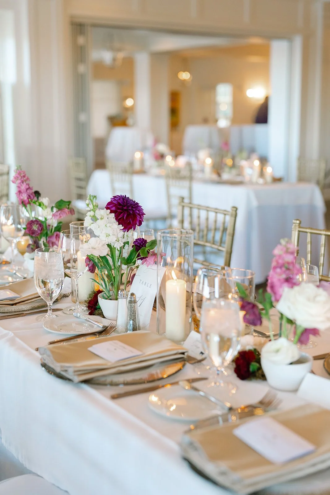 Elegant wedding reception table setting with floral centerpieces, candles, glassware, and folded napkins in a bright, decorated room.
