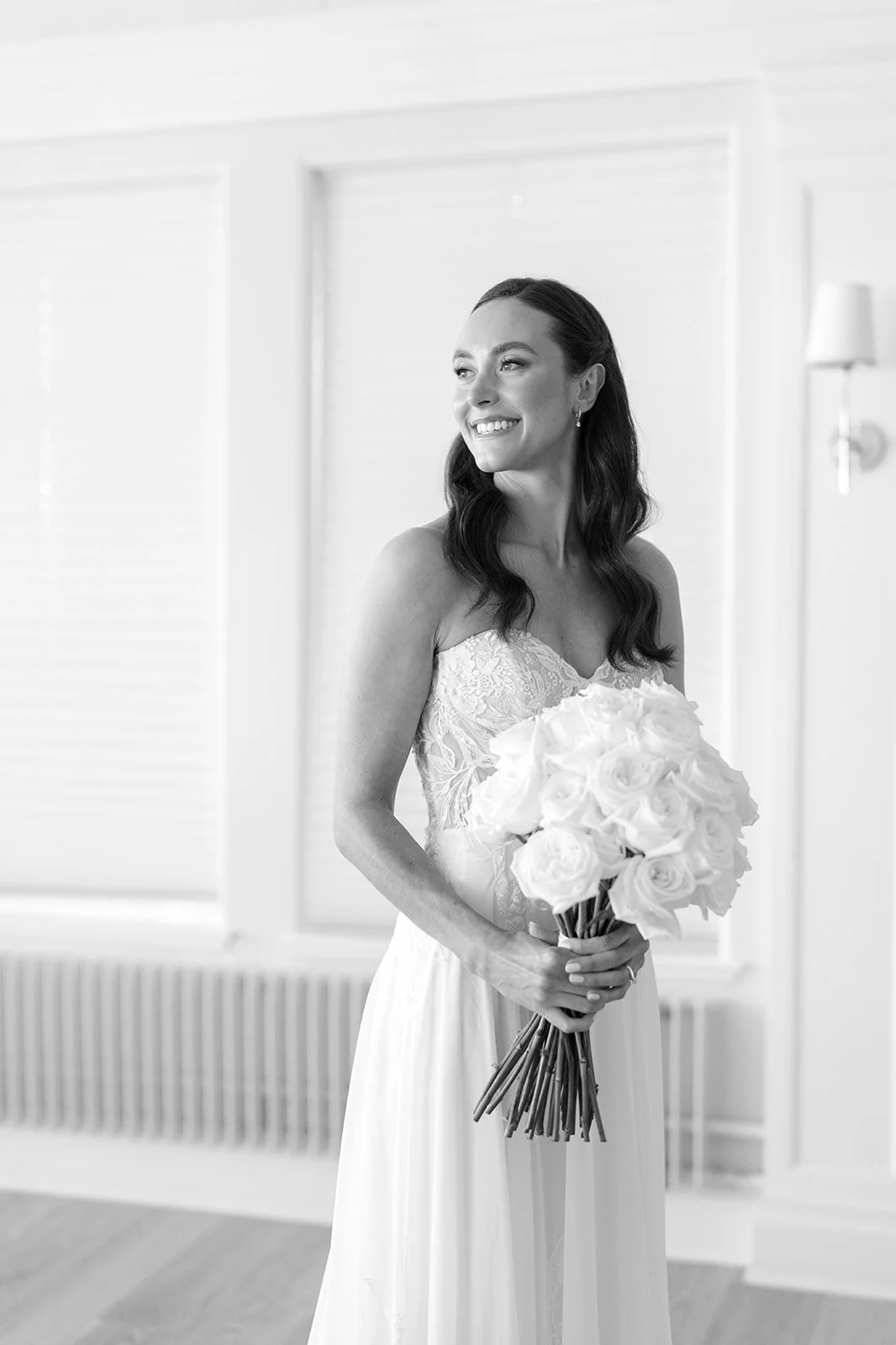 A smiling bride with long dark hair, wearing a lace wedding dress, holding a large bouquet of white roses in a bright room.
