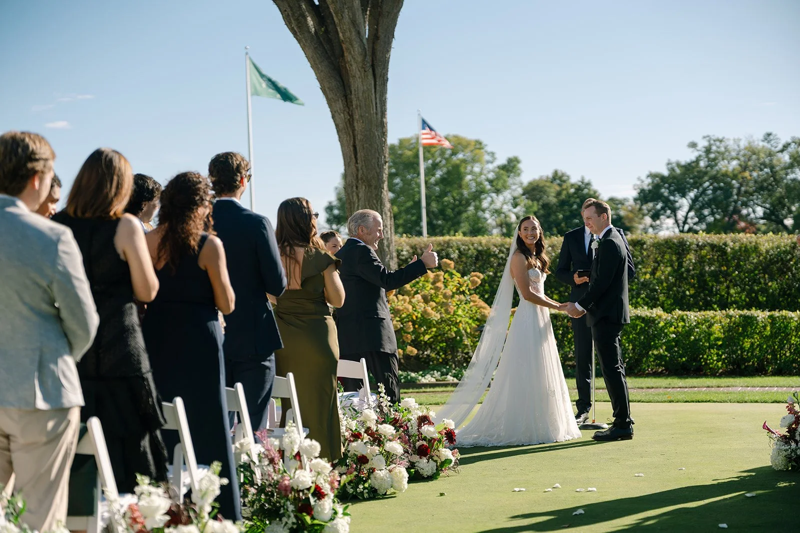 A wedding ceremony outdoors with a bride and groom holding hands, surrounded by friends and family, under a large tree with two flags in the background, on a sunny day with lush greenery.
