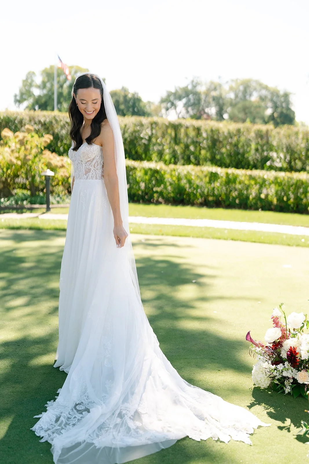 Bride in a white wedding dress standing on grass during the daytime, smiling and looking down, with a bouquet of flowers on the ground nearby. Minekahda club maroon mulberry florals wedding planning minneapolis wedding planner 