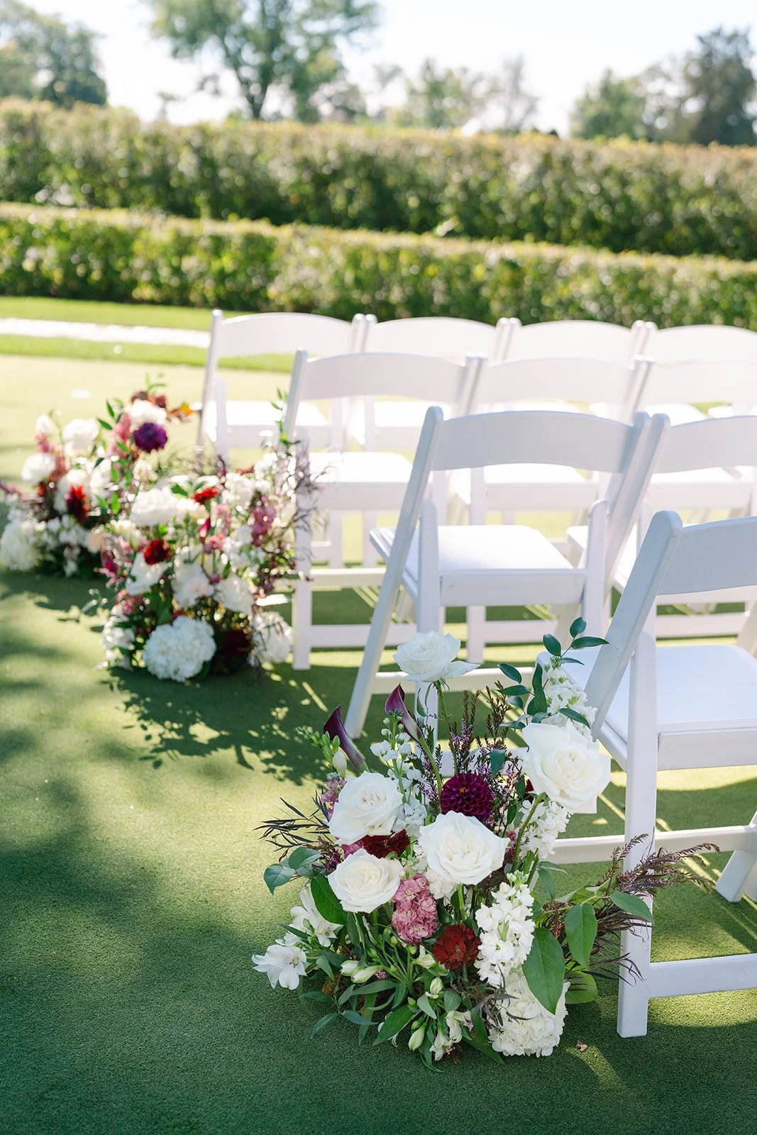 White chairs arranged in a row outdoors with colorful flower arrangements at the base, set against a green lawn and hedges on a sunny day.