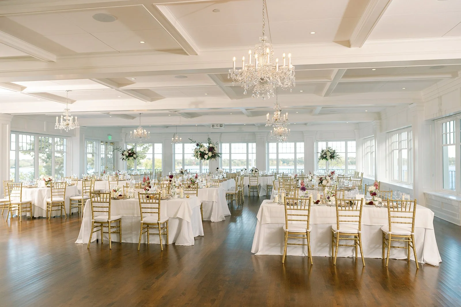 Elegant banquet hall with white tablecloths, gold chairs, floral centerpieces, large windows, and chandeliers hanging from a white ceiling.