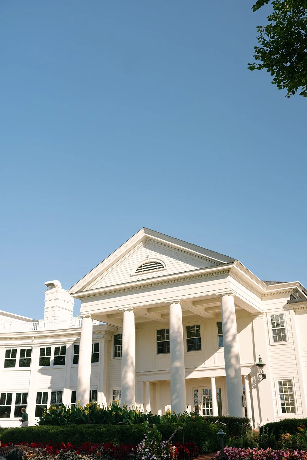 A large white house with columns, multiple windows, and a porch, surrounded by a garden with flowers and greenery, under a clear blue sky. minekahda club