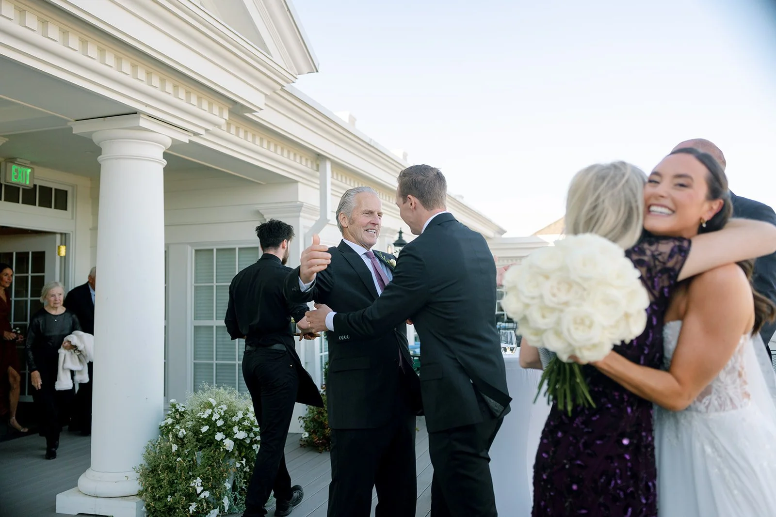 People celebrating at a wedding reception, with a woman in a white dress holding a bouquet of white roses hugging a woman in a dark purple dress and smiling.