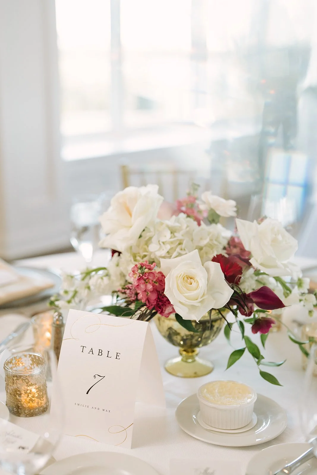 Elegant wedding table setup with a floral centerpiece of white and pink flowers, a table number card, and candle holders.