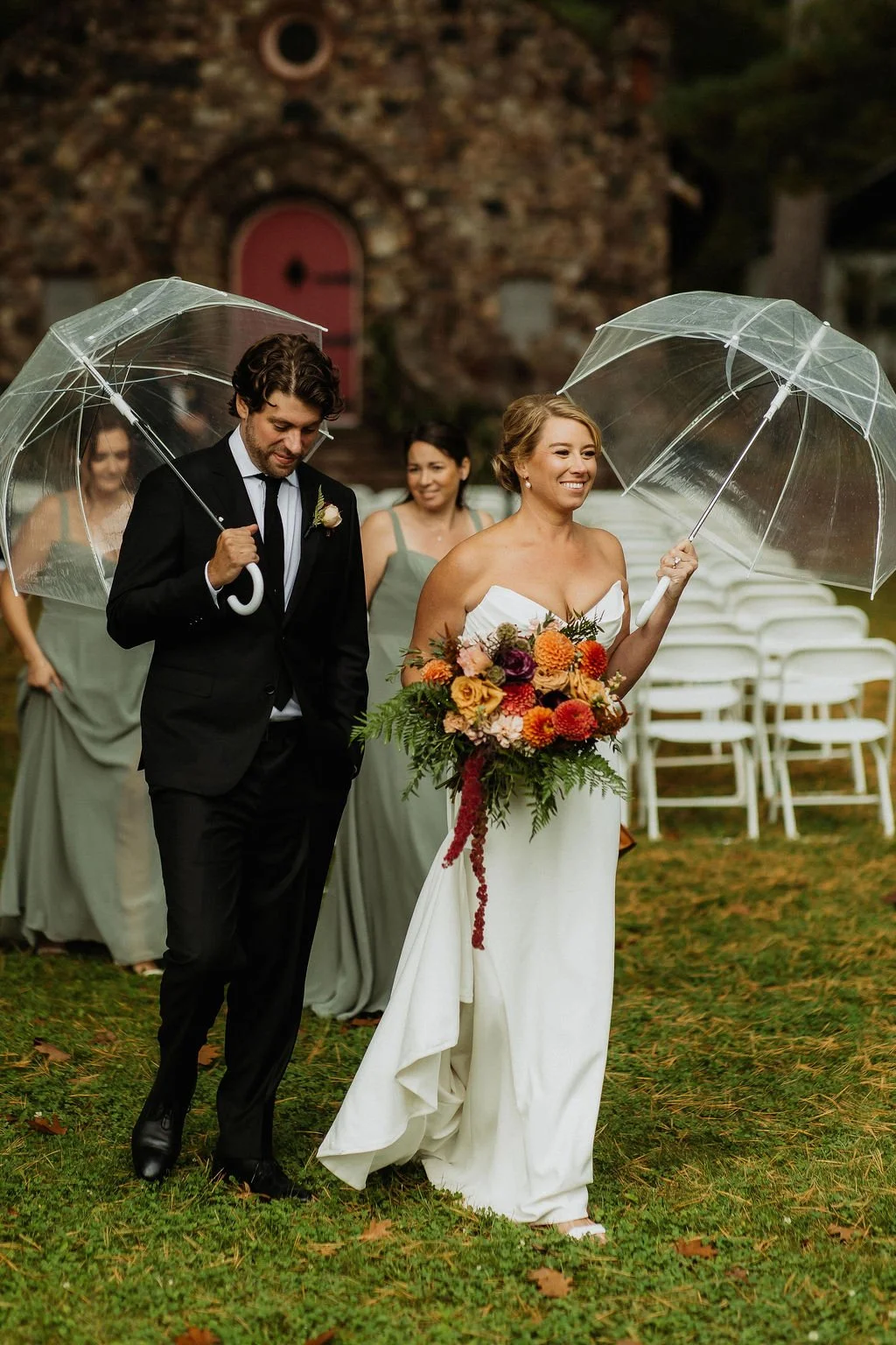 A bride and groom at an outdoor wedding, holding clear umbrellas, with smiling bridesmaids in green dresses in the background. camp foley wedding planning floral northern minnesota wedding jenkins nisswa