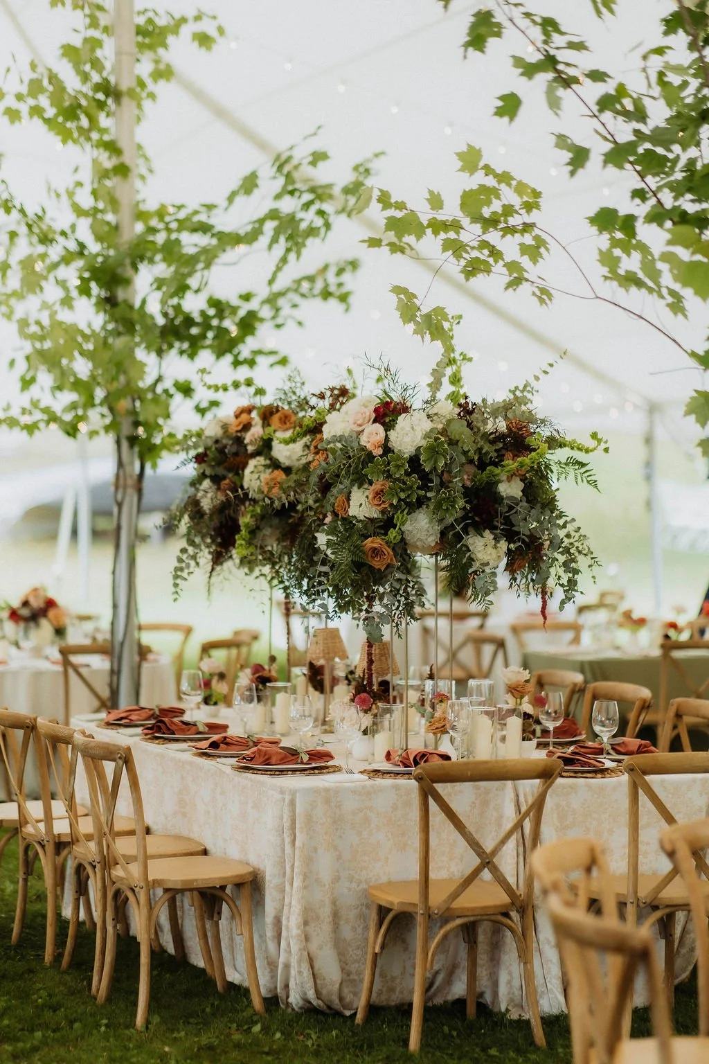 Wedding reception table with elegant floral centerpiece covered with white, peach, and dark red flowers and surrounded by wooden chairs, set outdoors under a tent with green leaves hanging from the ceiling.