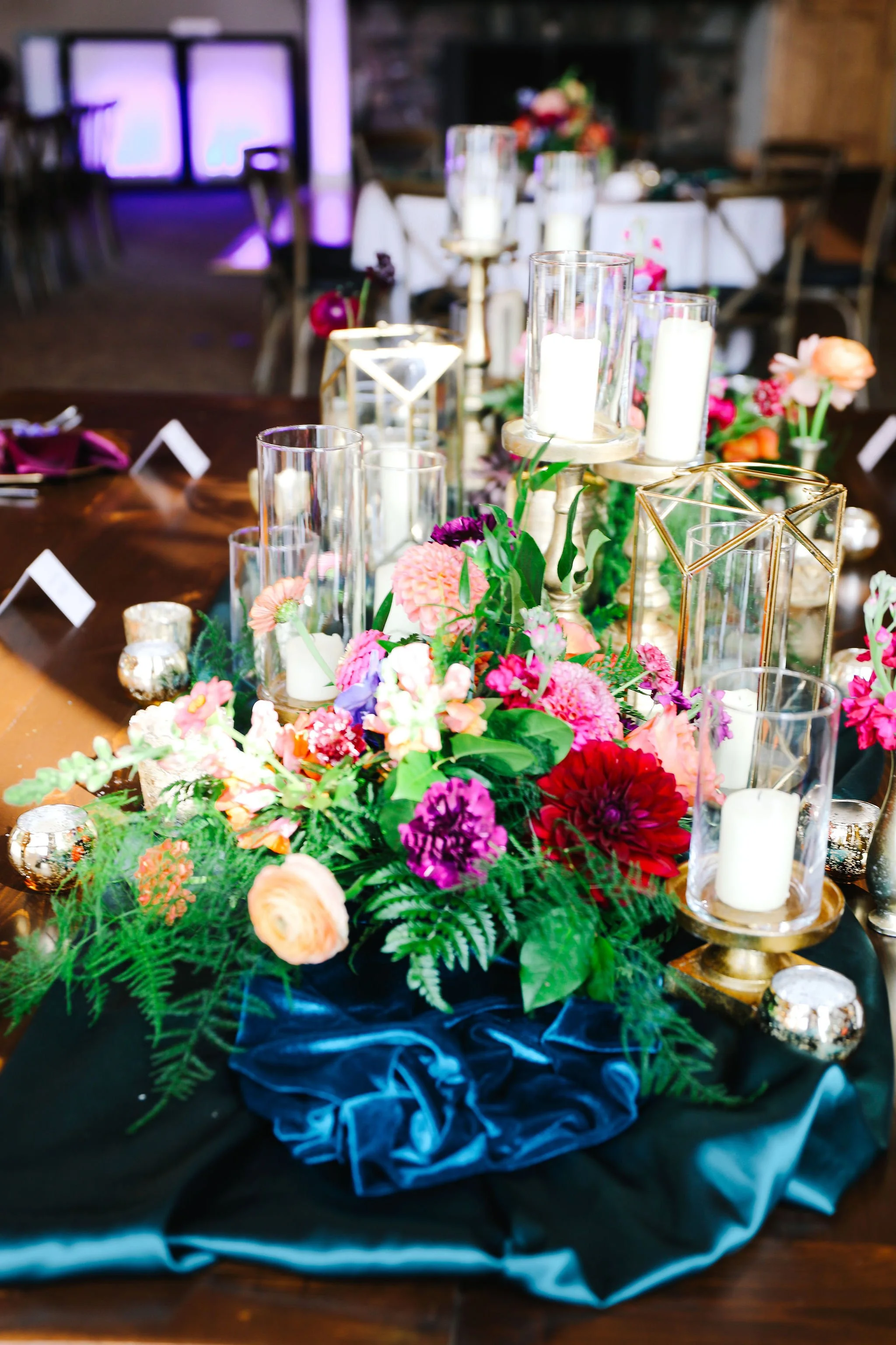 Decorative centerpiece with pink, purple, and red flowers, surrounded by white candles in glass holders, gold geometric accents, and a dark blue fabric runner on a wooden table at an event. jewel tone