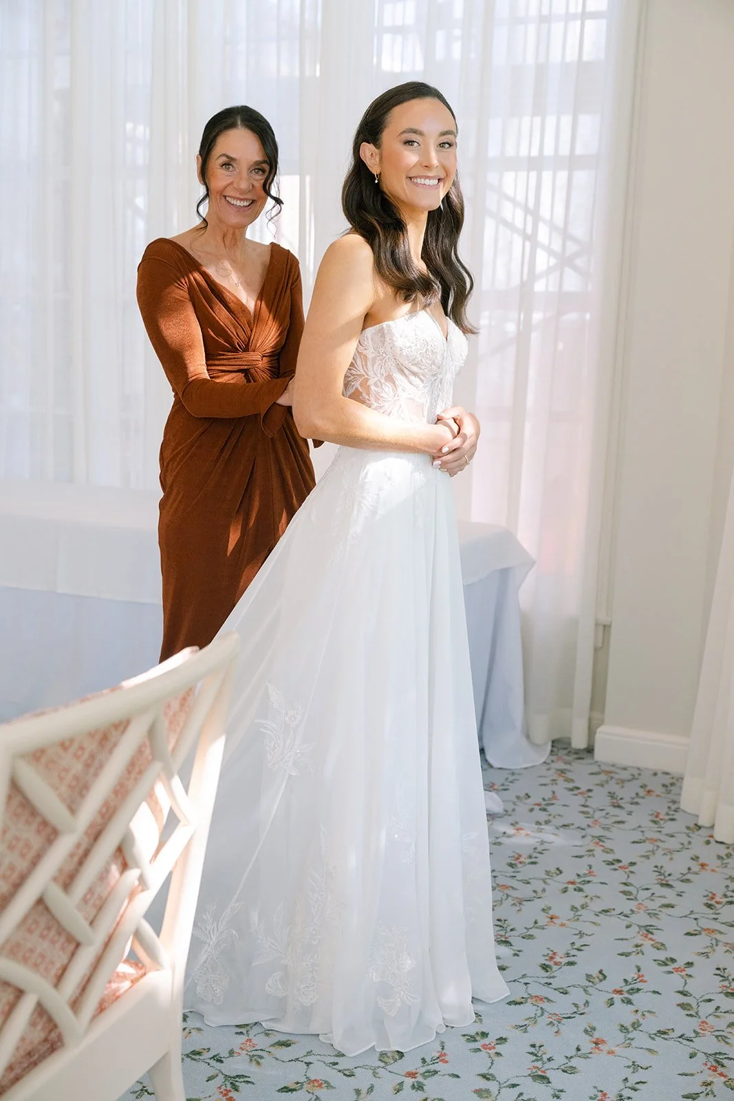 A woman in a white wedding dress smiling as she is helped by another woman in a brown dress, both standing inside a room with white curtains and floral carpet.Minekahda club maroon mulberry florals wedding planning minneapolis wedding planner 