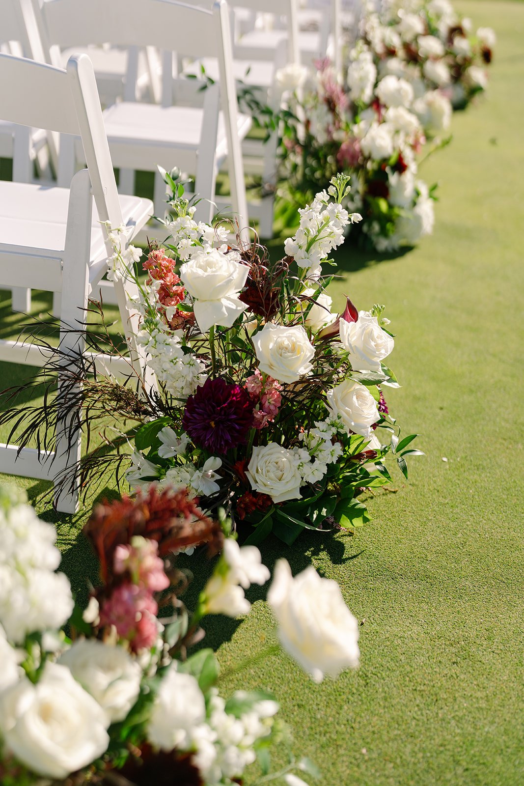 White chairs in a row on a green lawn with floral arrangements along the aisle for a wedding ceremony. Outdoor ceremony Minekahda club maroon mulberry florals wedding planning minneapolis wedding planner. wedding aisle flowers