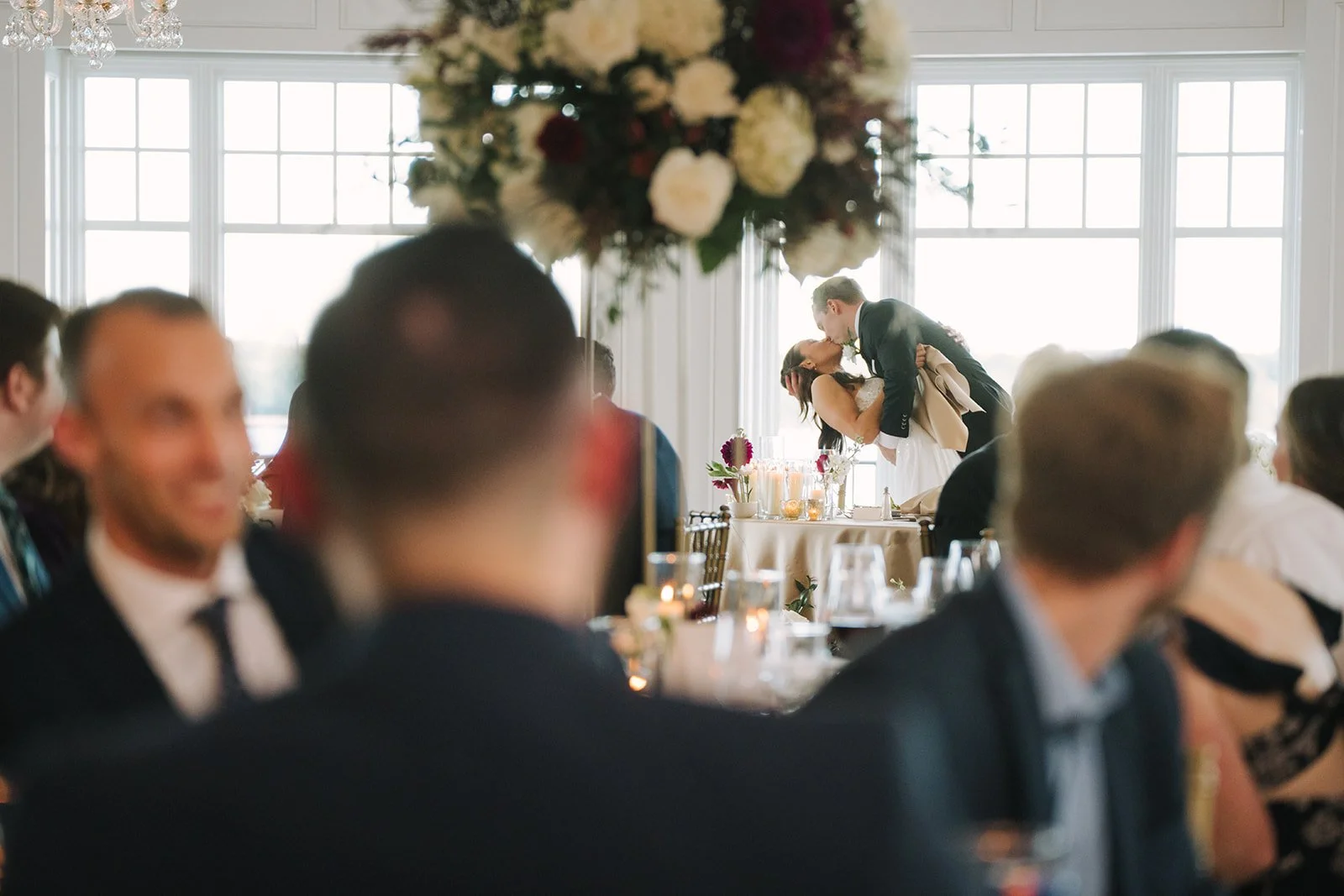 A wedding reception with a bride and groom sharing a kiss in the background at their head table, decorated with candles and flowers, while guests seated at tables in foreground watch.