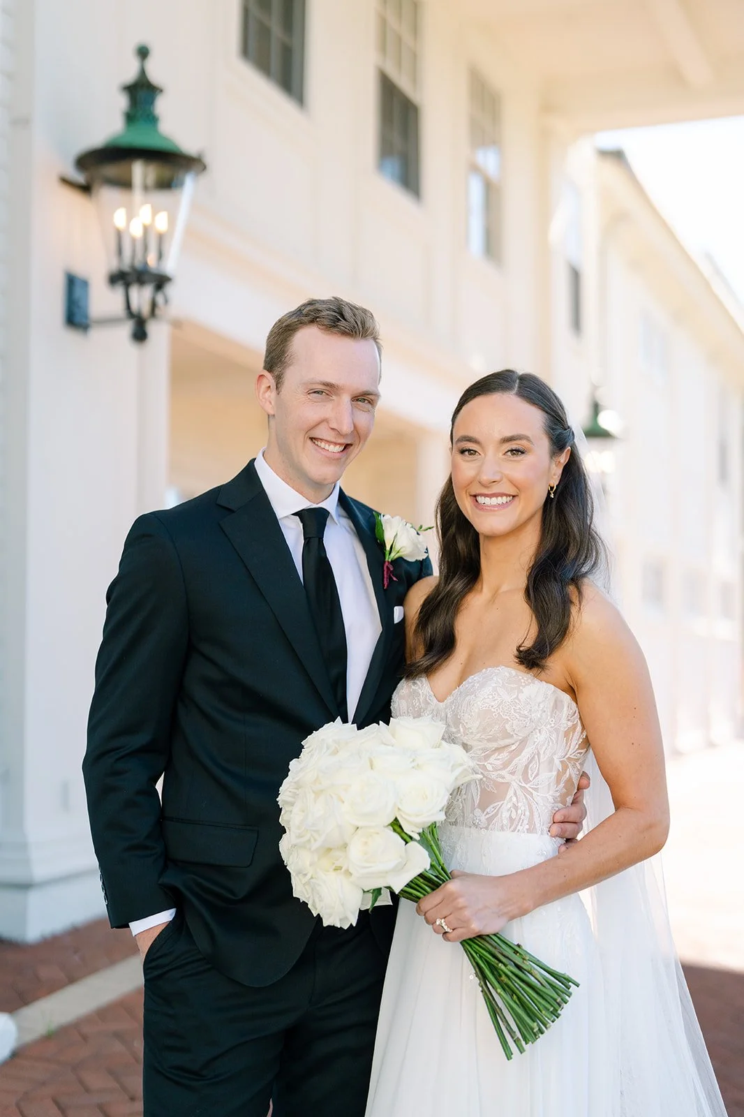 A bride and groom smiling at the camera outside a wedding venue, with the bride holding a bouquet of white roses.