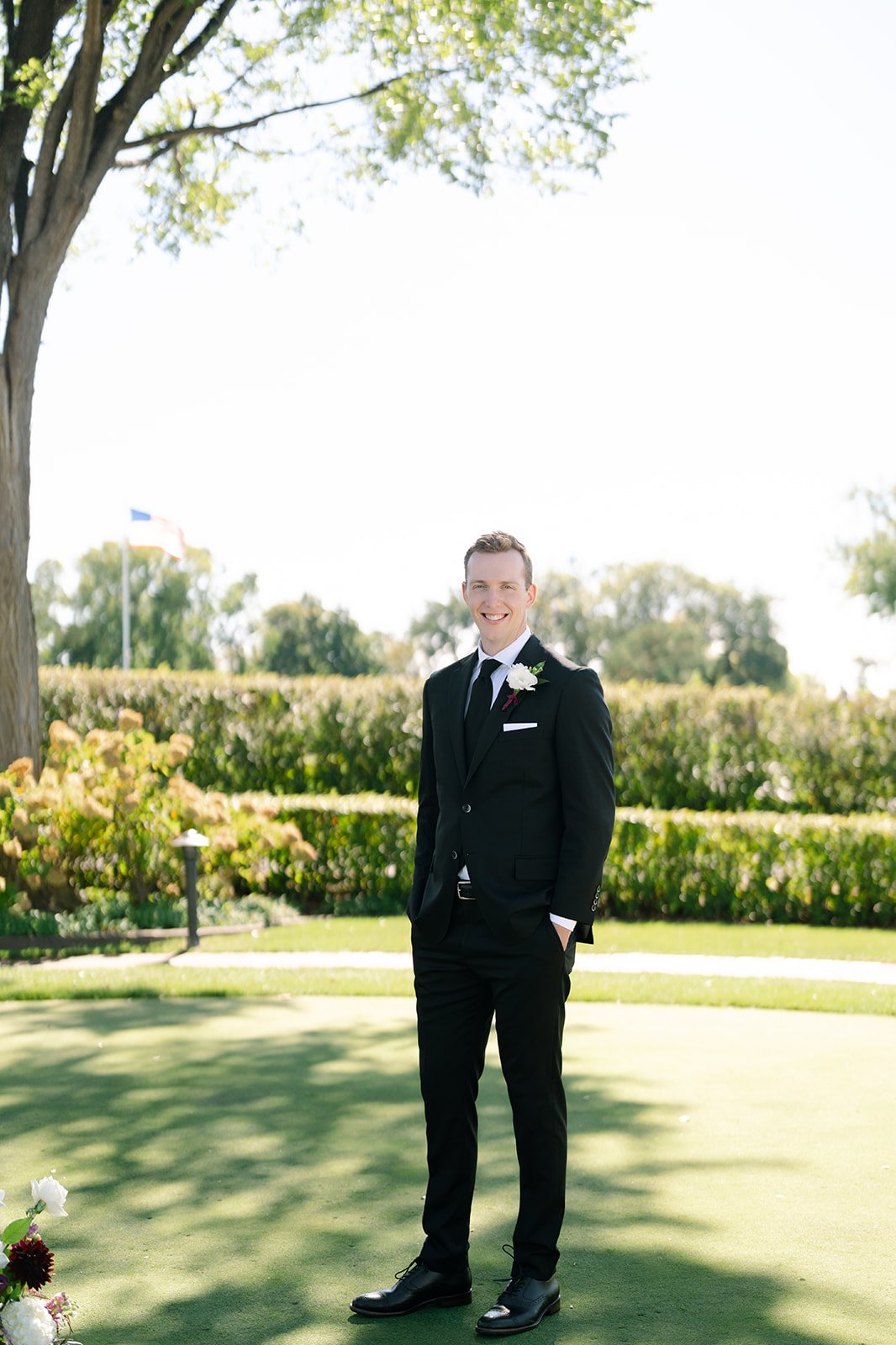 A man in a black suit and tie, with a white shirt, standing outdoors on a sunny day, smiling, with green trees and bushes in the background.