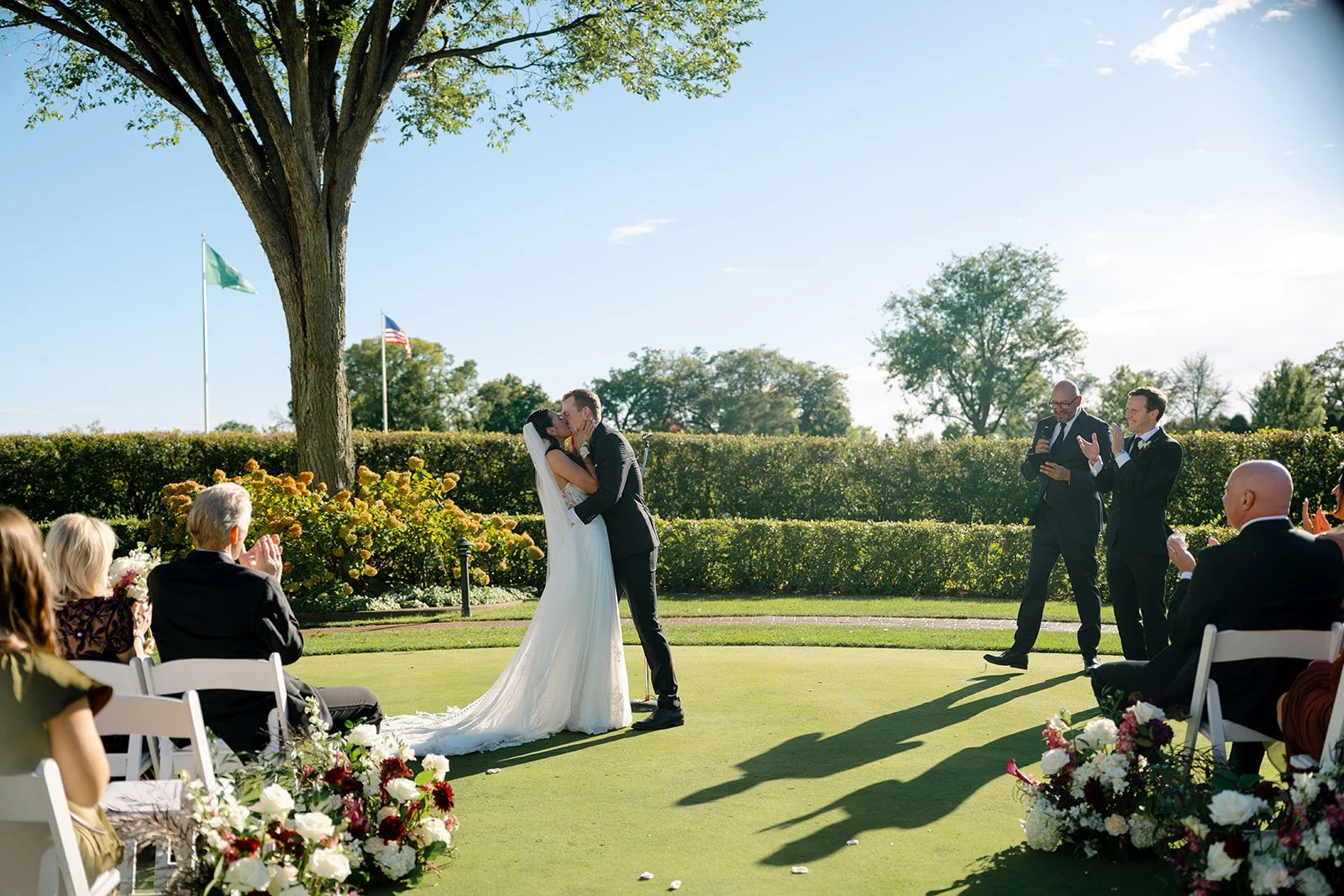 A wedding ceremony outdoors with a bride and groom kissing under a tree, surrounded by guests seated and clapping, with flags in the background on a sunny day.