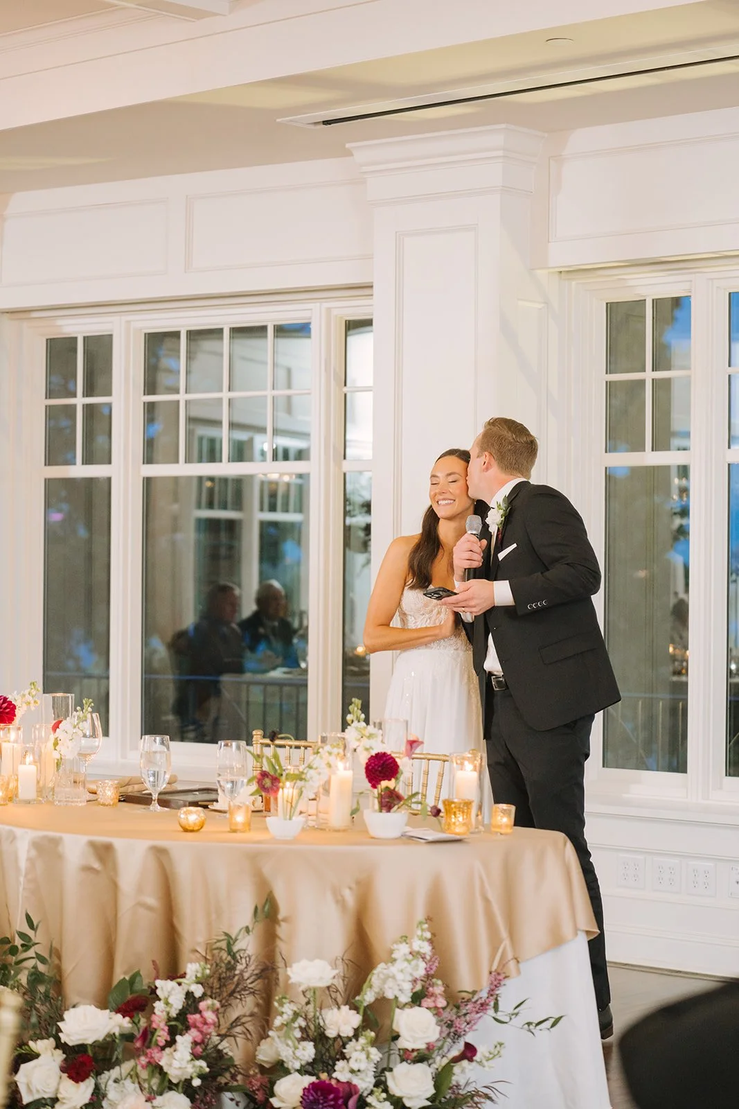 Bride and groom giving a speech at their wedding reception, with the groom kissing the bride on the cheek, standing by a decorated table with flowers and candles in a bright room with large windows.