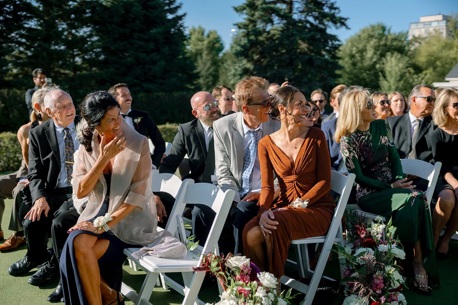 Attendees at a wedding ceremony outdoors, seated on white chairs, laughing and smiling in sunlight, with green trees and buildings in the background.