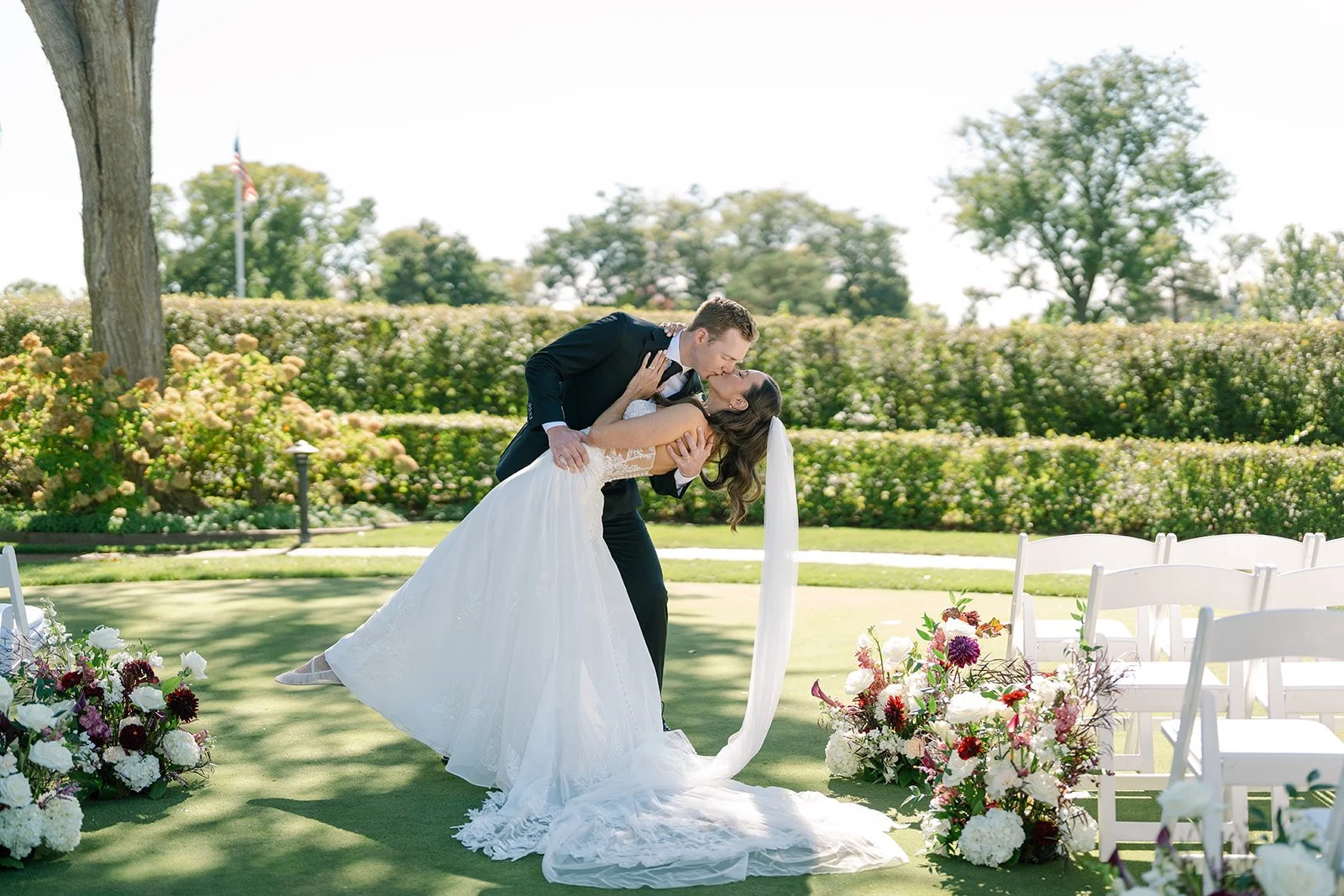 A bride and groom share a kiss outdoors at their wedding ceremony, with chairs and floral arrangements surrounding them on a grassy area under trees.