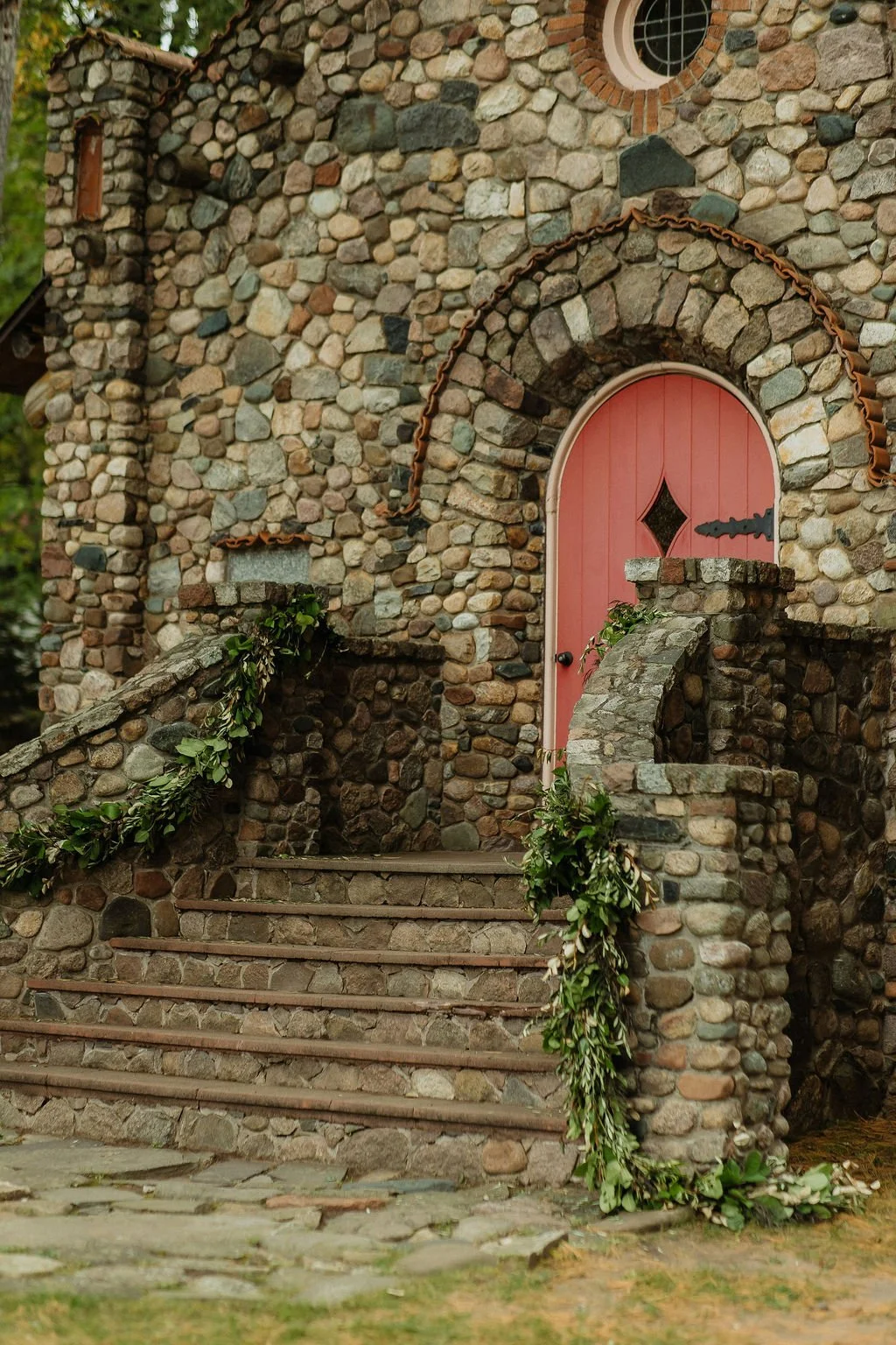 A stone house with a curved, pink wooden door and circular window above. The house features a stone staircase and handrails decorated with green foliage. camp foley wedding planning floral northern minnesota wedding jenkins nisswa