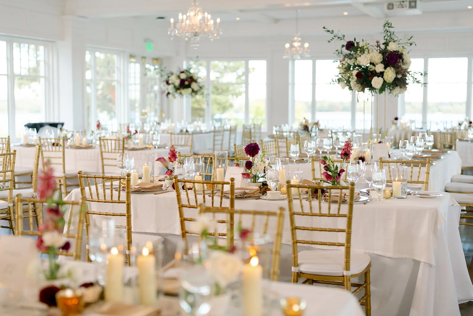 Elegant event space with round tables covered in white tablecloths, decorated with pink and white flowers, candles, and glassware. The room has large windows, gold chairs, and a chandelier, with a view of water and trees outside.