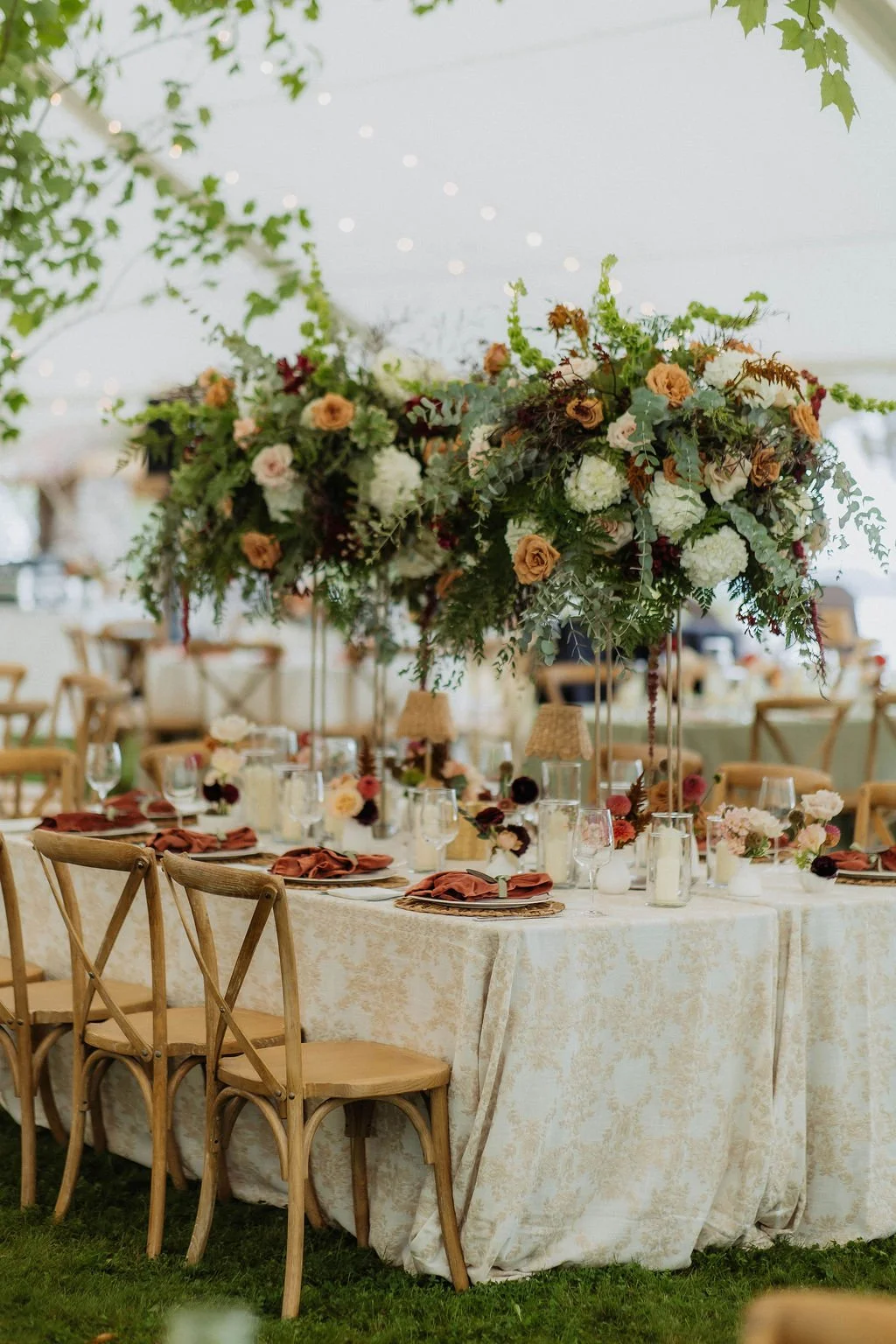 Wedding reception table decorated with floral centerpieces, set with plates, glasses, and napkins in a tented outdoor setting. tall florals on harlow stands wedding camp foley