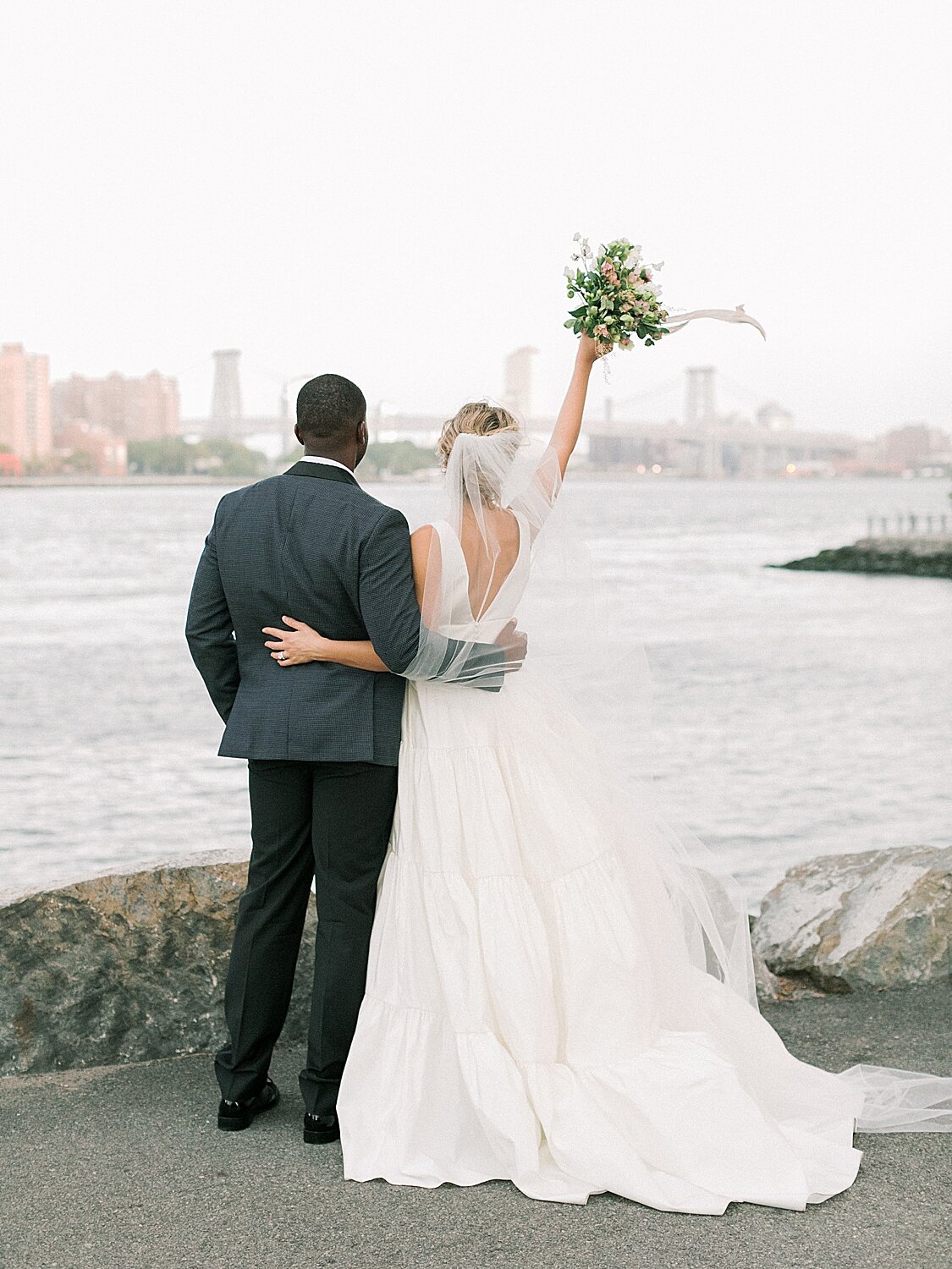 bride cheers with bouquet while standing facing Hudson River with groom | Asher Gardner Photography | Intimate Ceremony in DUMBO New York