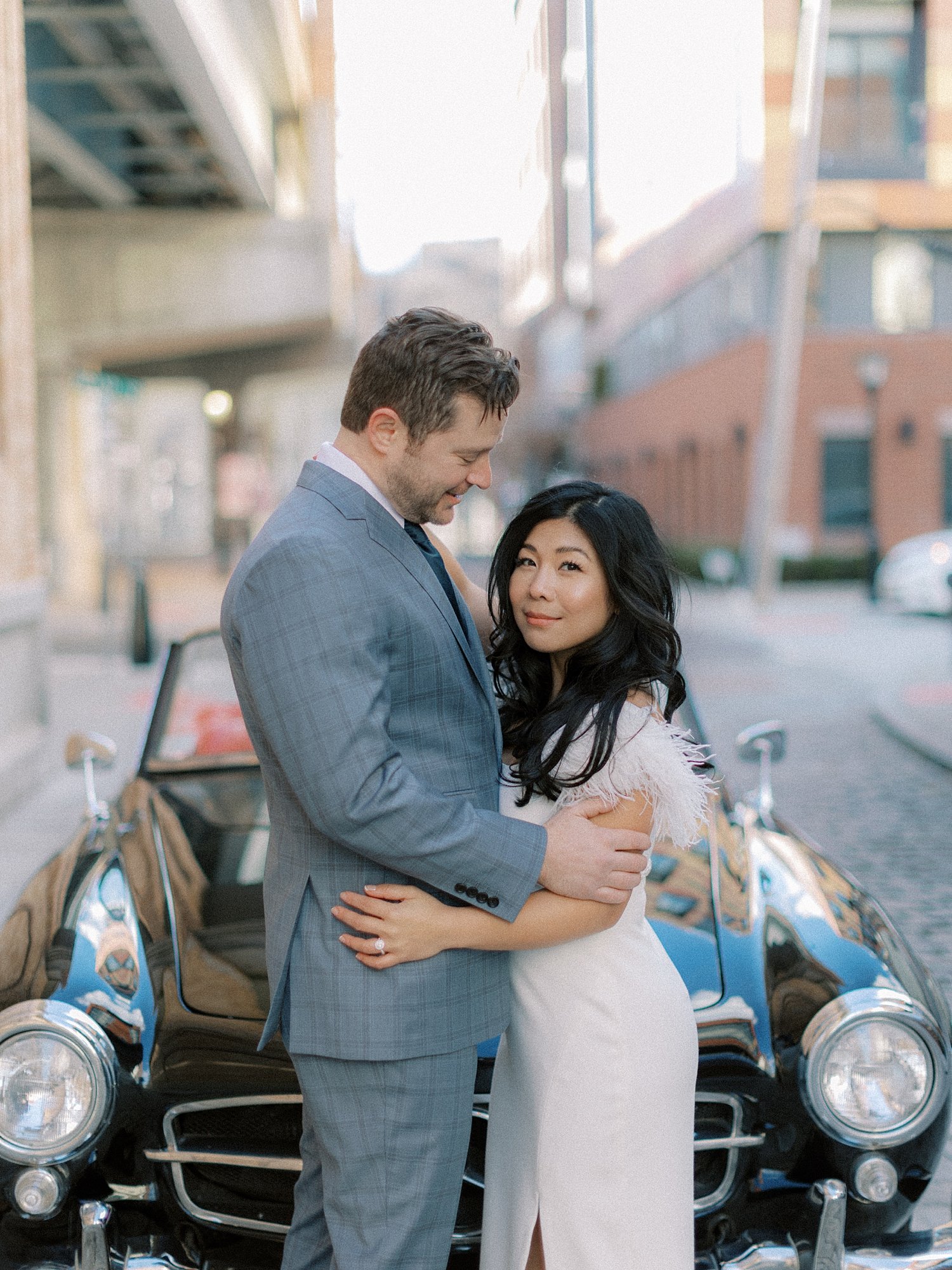 man in blue suit hugs woman in chic white dress in front of black classic Mercedes Benz 190sl