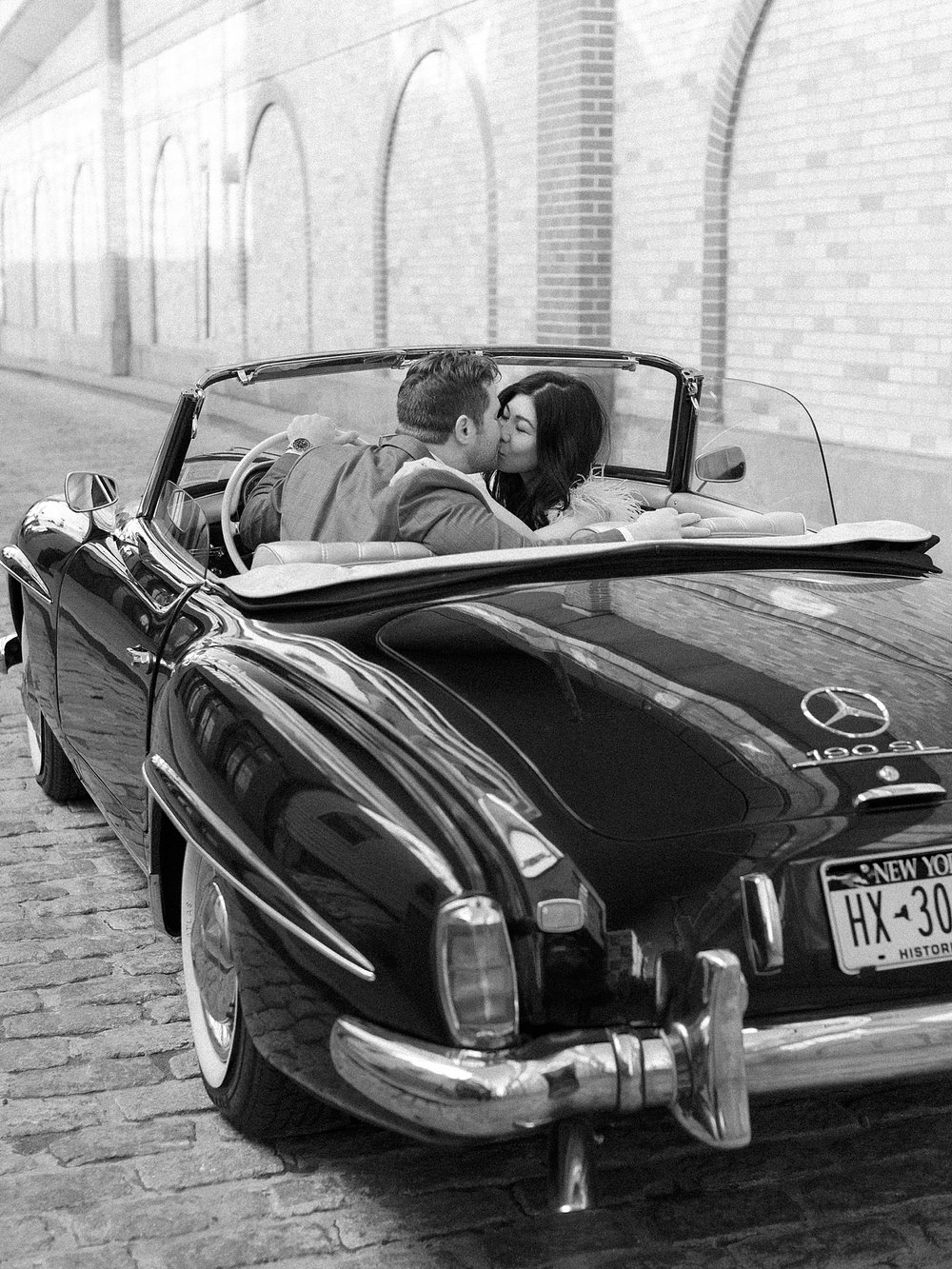 engaged couple sits in classic Mercedes Benz 190sl leaning for a kiss on street near Carpe Diem in New Jersey